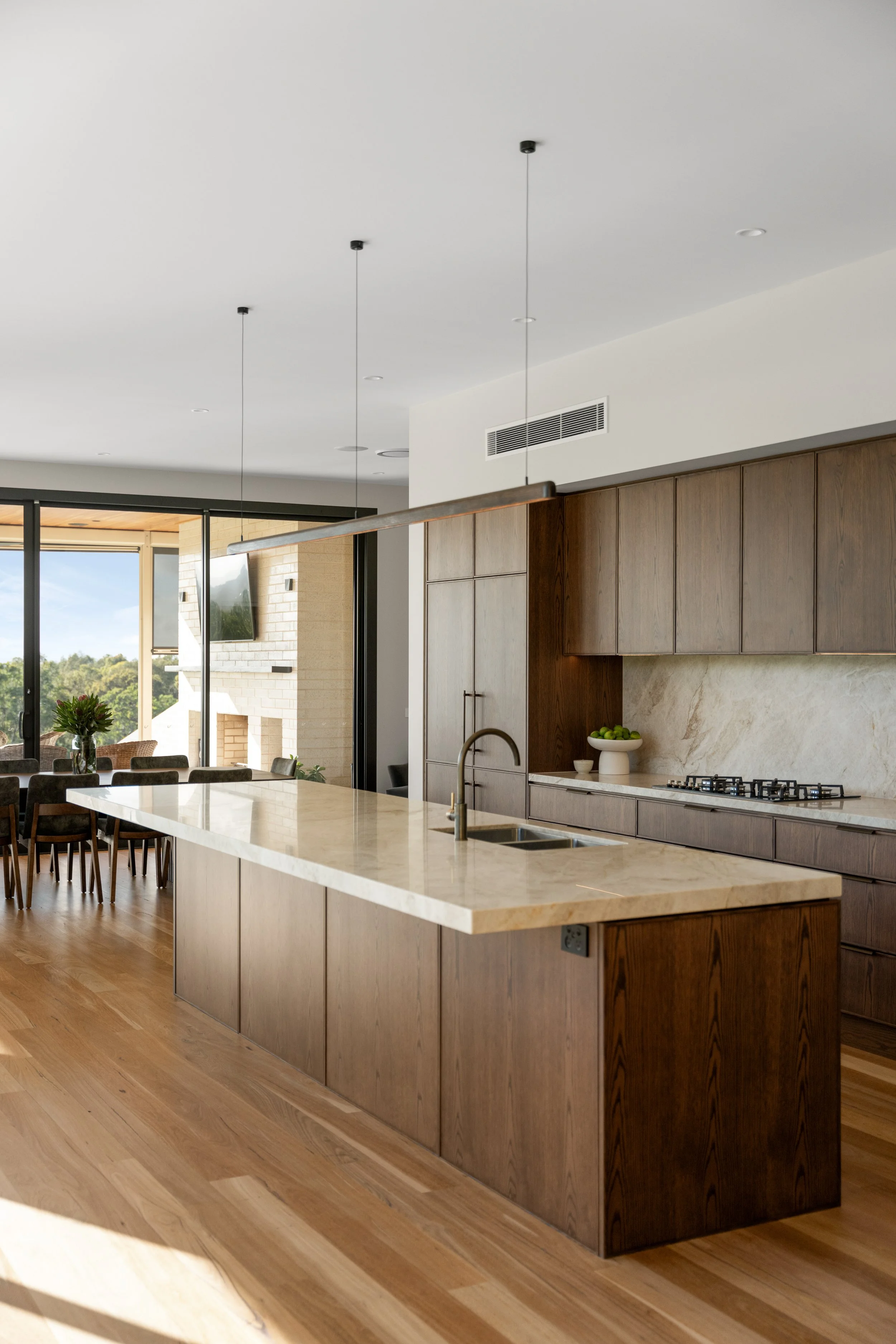 Modern kitchen with large marble-topped island, wooden cabinets, and a view of a dining area with chairs and a window showing trees outside.