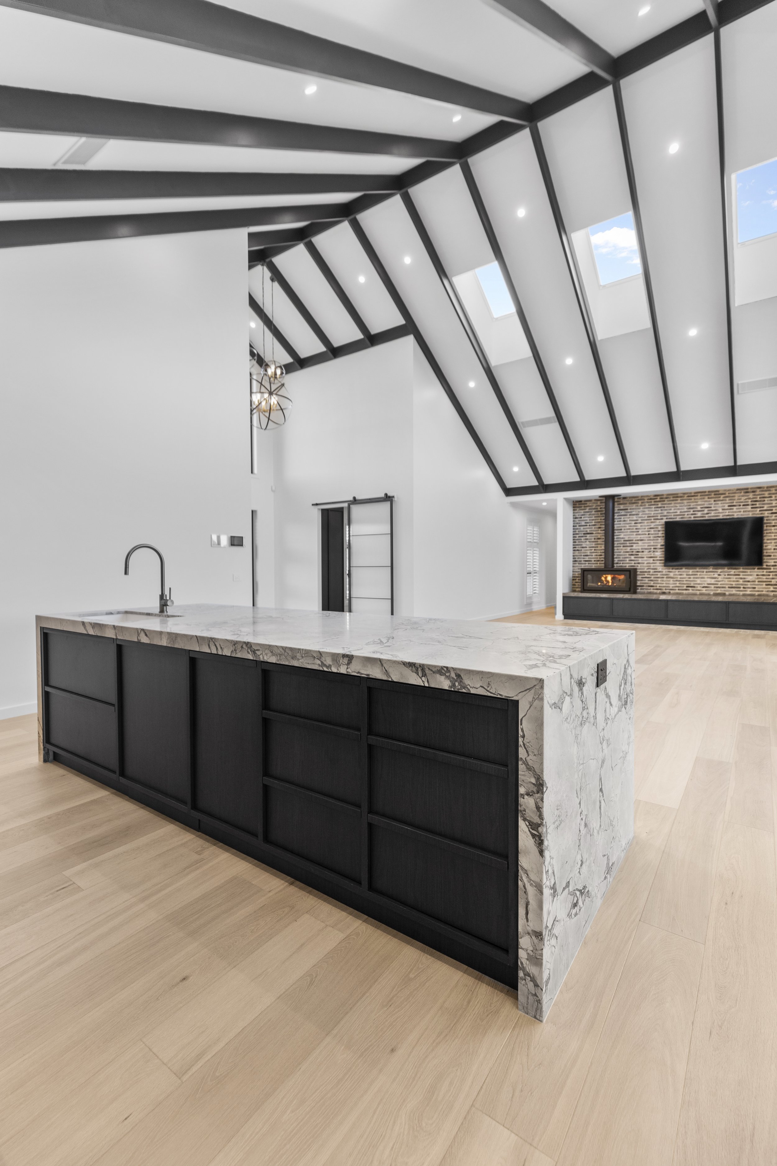 Modern open-concept kitchen and living room with vaulted ceiling, skylights, white walls, light wood flooring, black kitchen island with marble countertop, fireplace, and flat-screen TV.