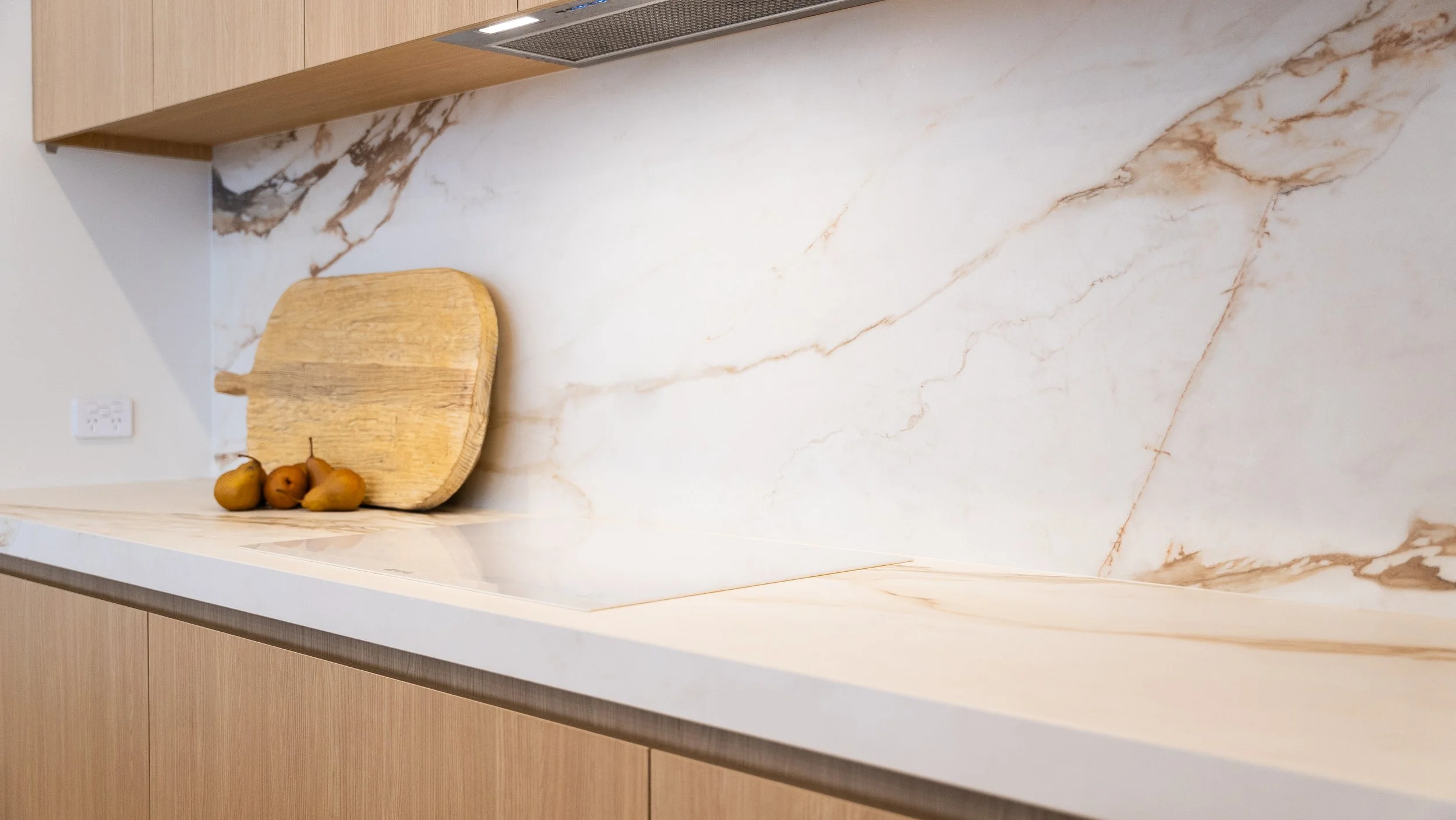 A kitchen countertop with a cream-colored marble surface featuring brown veins, a wooden cutting board, and a small bunch of pears.