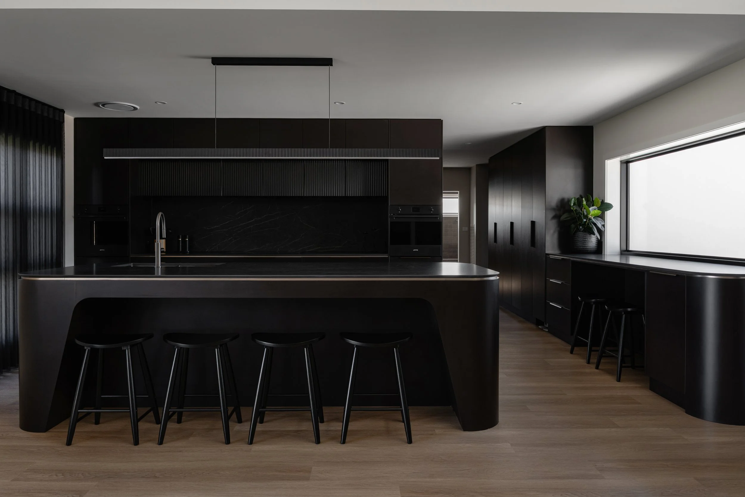 Modern kitchen with a large dark island, black stools, black cabinets, and a large window with a potted plant near it.