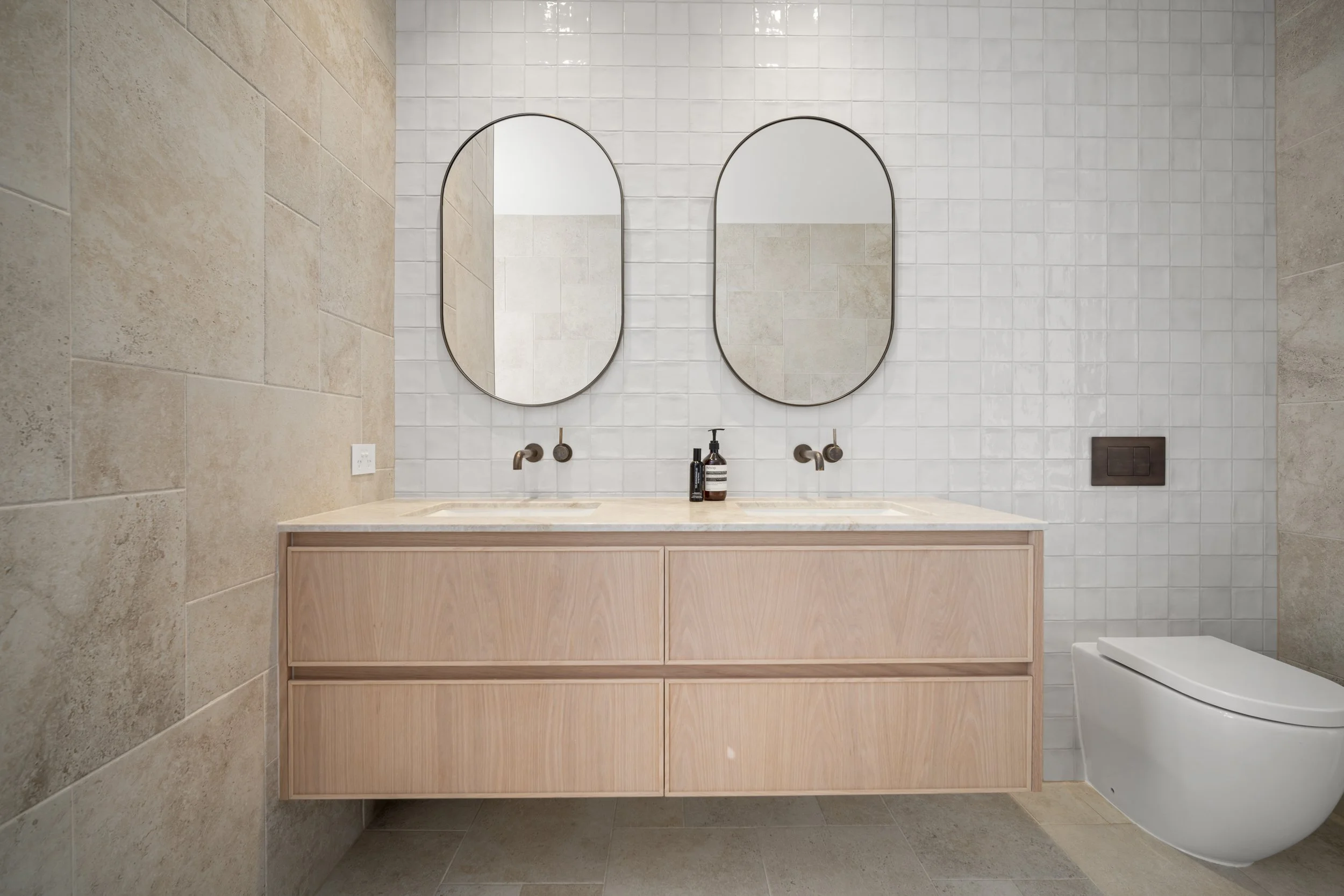 Modern bathroom with a wooden vanity, two oval mirrors, and a white toilet, all featuring neutral tones and minimal decor.