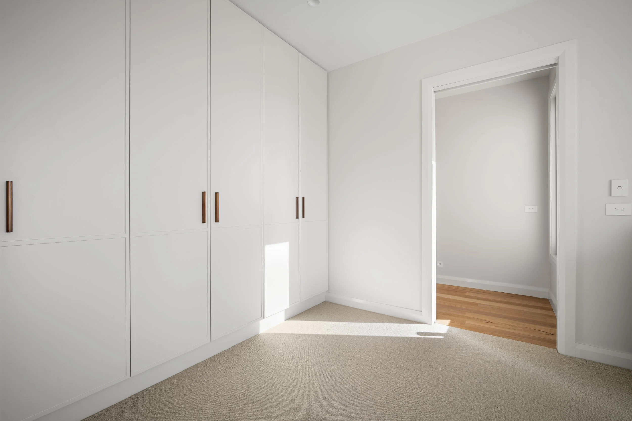 A white built-in closet with multiple cabinets and wooden handles in a minimalist room. Part of an open doorway revealing a room with wooden flooring and white walls.