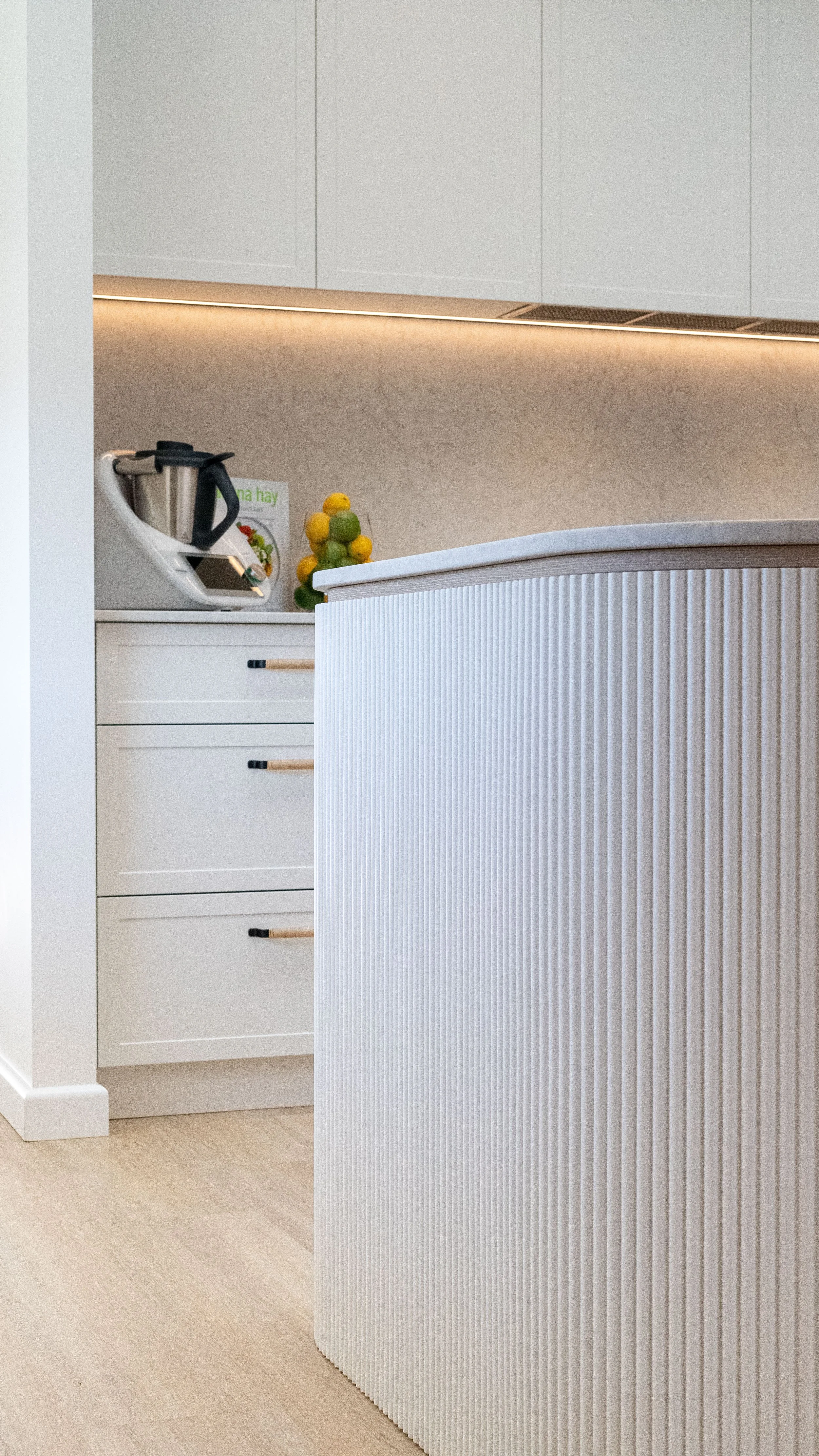 Close-up of a modern kitchen with white cabinetry, a textured white kitchen island, and a countertop with a blender, a small book, and a decorative fruit sculpture.