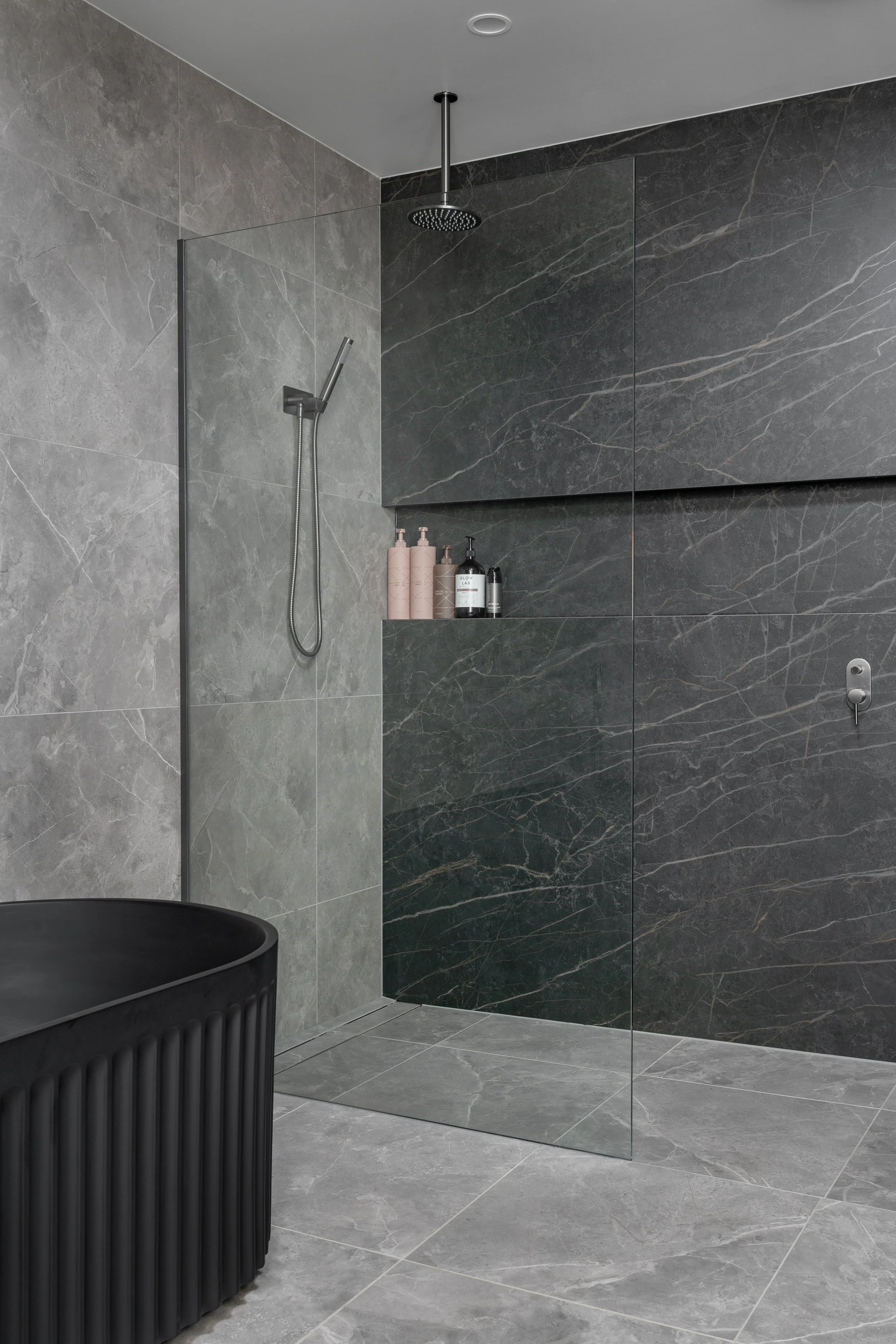 Modern shower area with gray marble tiles, a rainfall showerhead, a handheld shower, and a small shelf with bottles, separated from the rest of the bathroom by a glass partition.