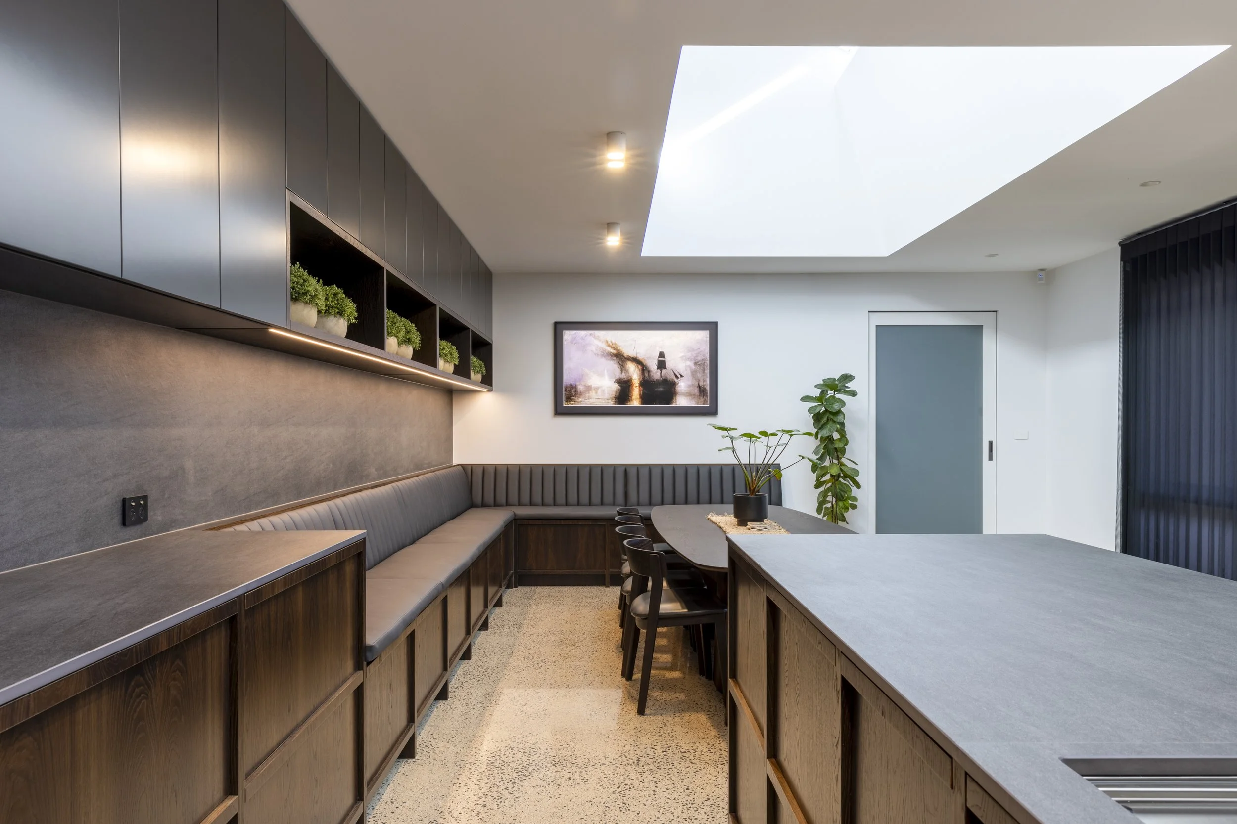 Modern dining area with built-in seating, wooden cabinetry, and a large table with black chairs. Decorative plants on shelves, a framed painting on the wall, and a ceiling skylight providing natural light.