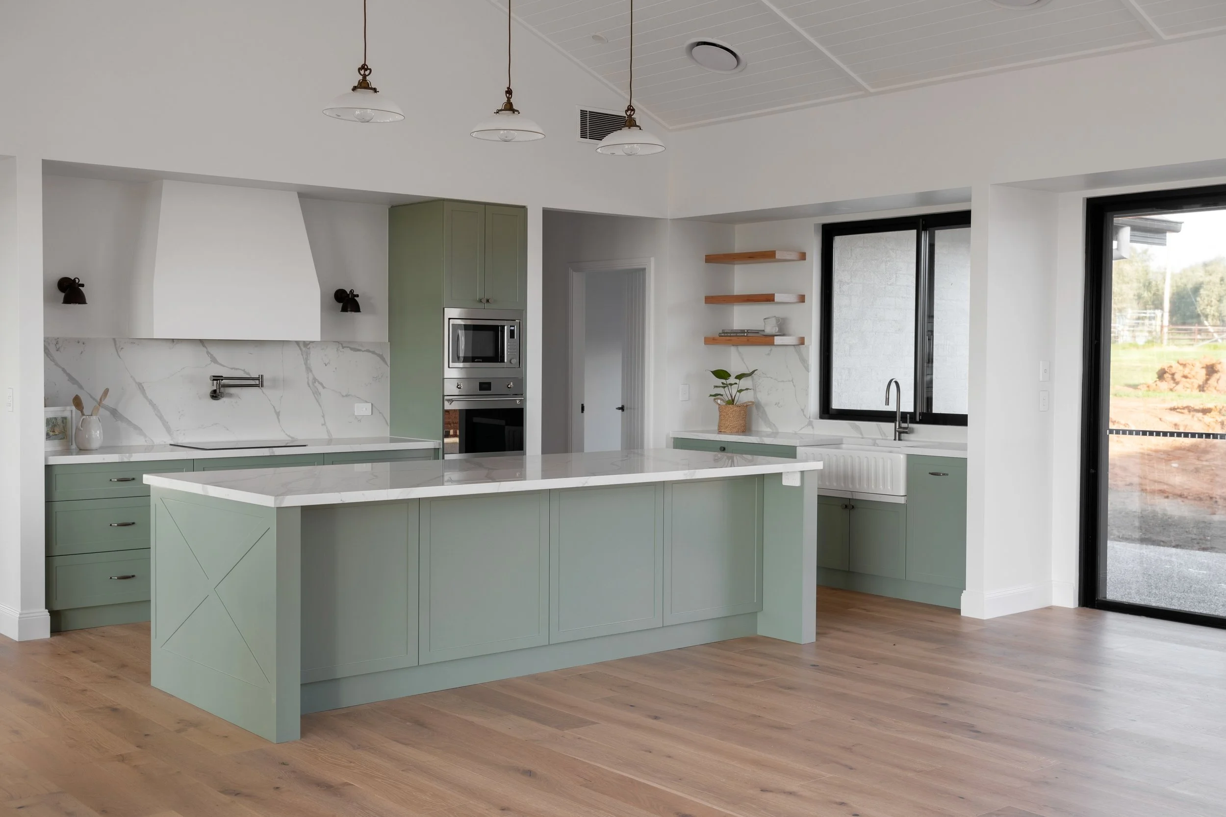 Modern kitchen with green cabinets, white marble countertops, a kitchen island, built-in microwave and oven, large windows, and hardwood flooring.