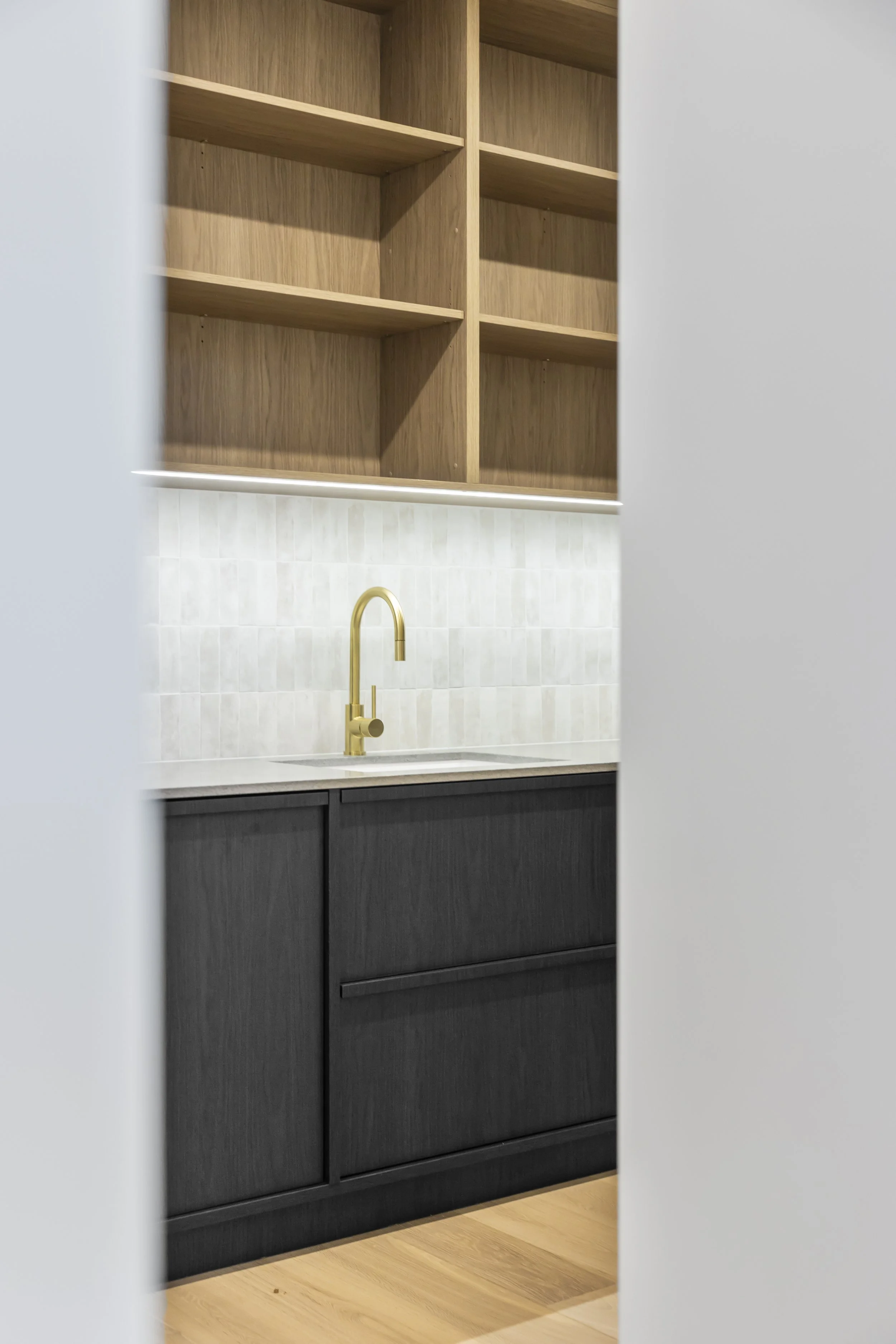 View through a partially opened white door showing a modern kitchen with black lower cabinets, a white countertop, a gold faucet, a wooden open shelf above the sink, white tile backsplash, and light wooden flooring.