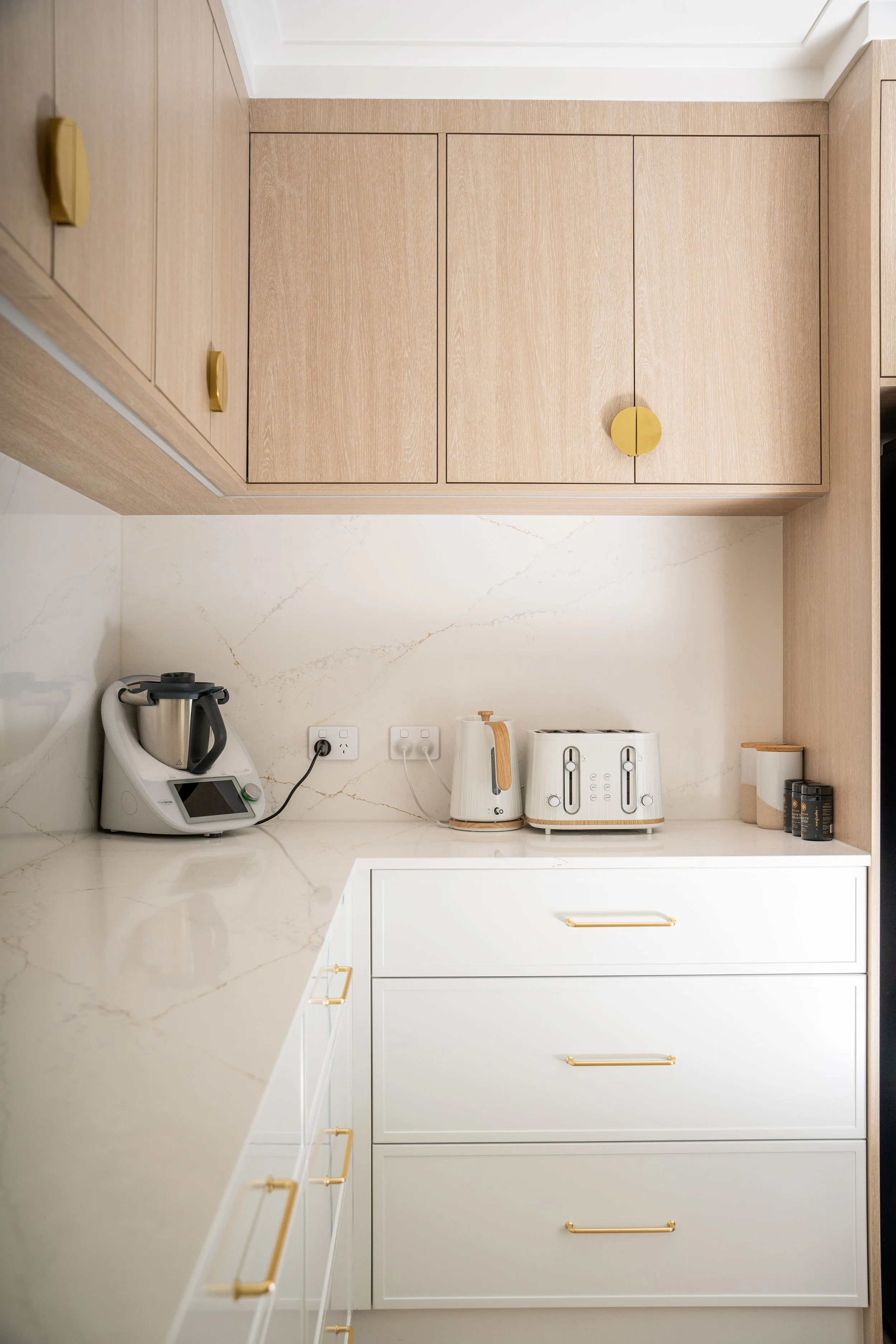 A modern kitchen countertop area with a white marble surface, featuring a Thermomix appliance on the left and a white toaster with wooden accents in the middle. Above, there are wooden cabinets with gold handles.