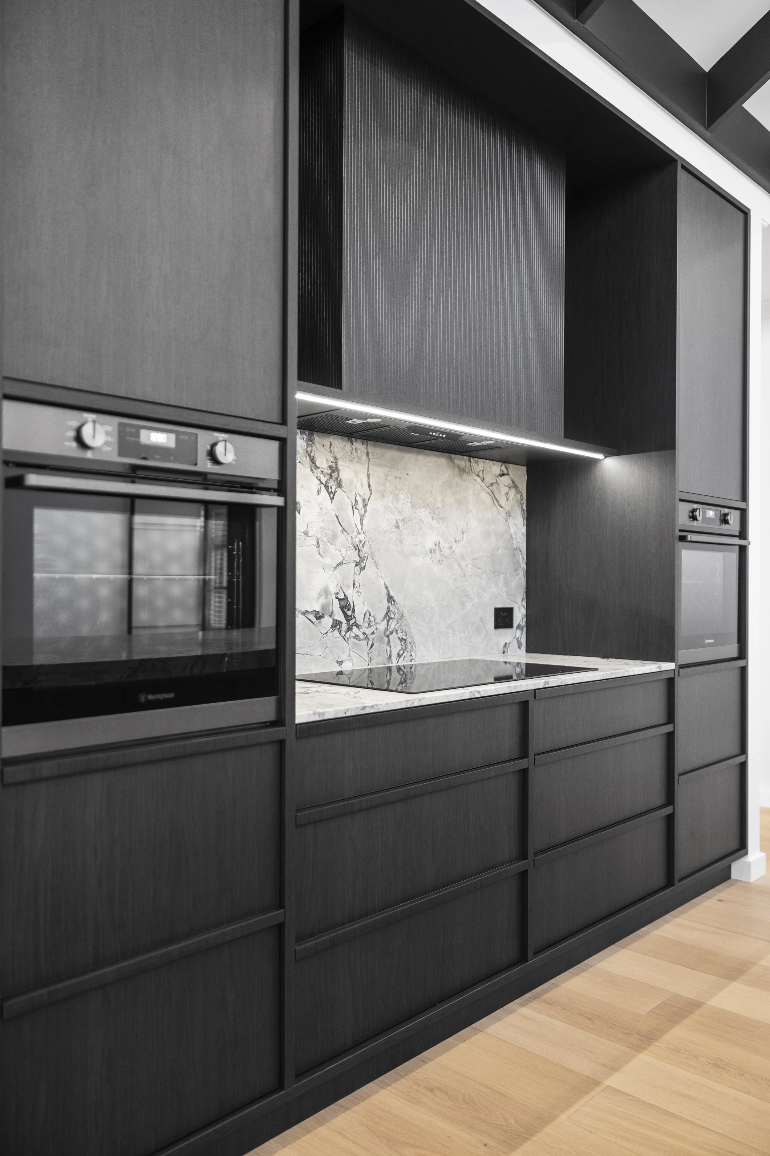 Modern kitchen with black cabinetry, marble backsplash, and built-in oven and microwave, with light wood flooring.