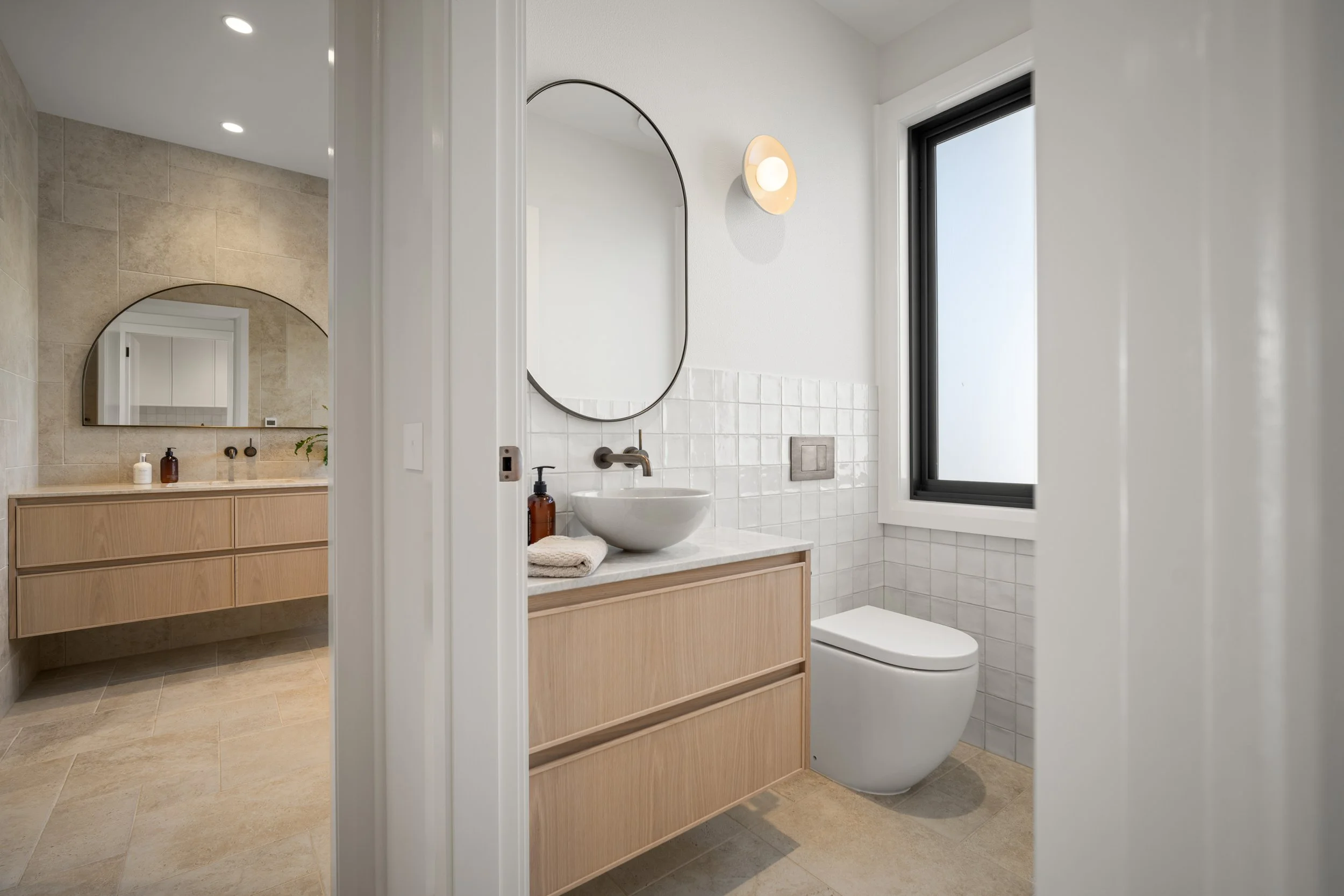 Modern bathroom with white walls, a round mirror above a vessel sink, a wooden vanity, a ceramic toilet, a black-framed window, and minimalist decor