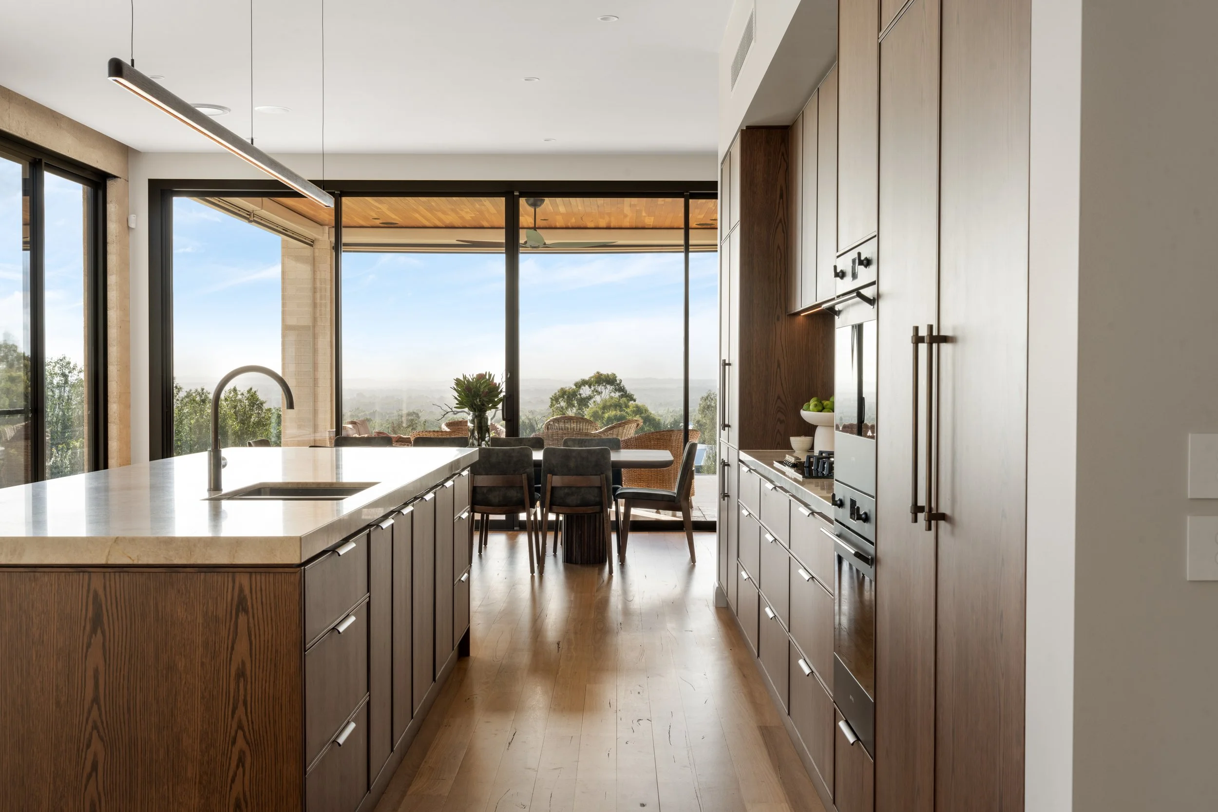 Modern kitchen with wooden cabinets, a large island with a sink, and large windows offering a view of trees and a blue sky.