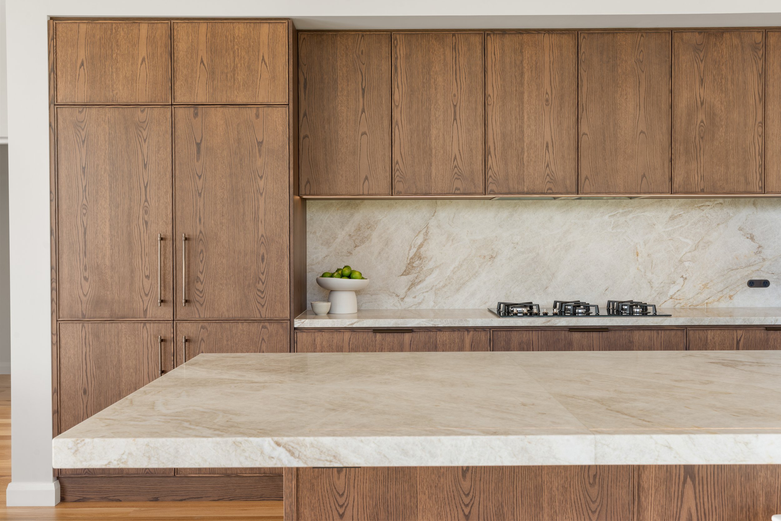 Modern kitchen with light-colored marble countertops, wooden cabinets, and a stove with two burners. There is also a white bowl of green limes on the counter.