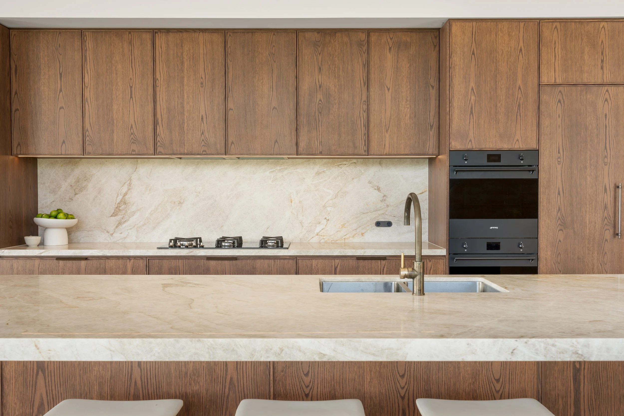 Modern kitchen with wooden cabinets, marble backsplash, and a marble island with a sink. There are three white chairs in front of the island.