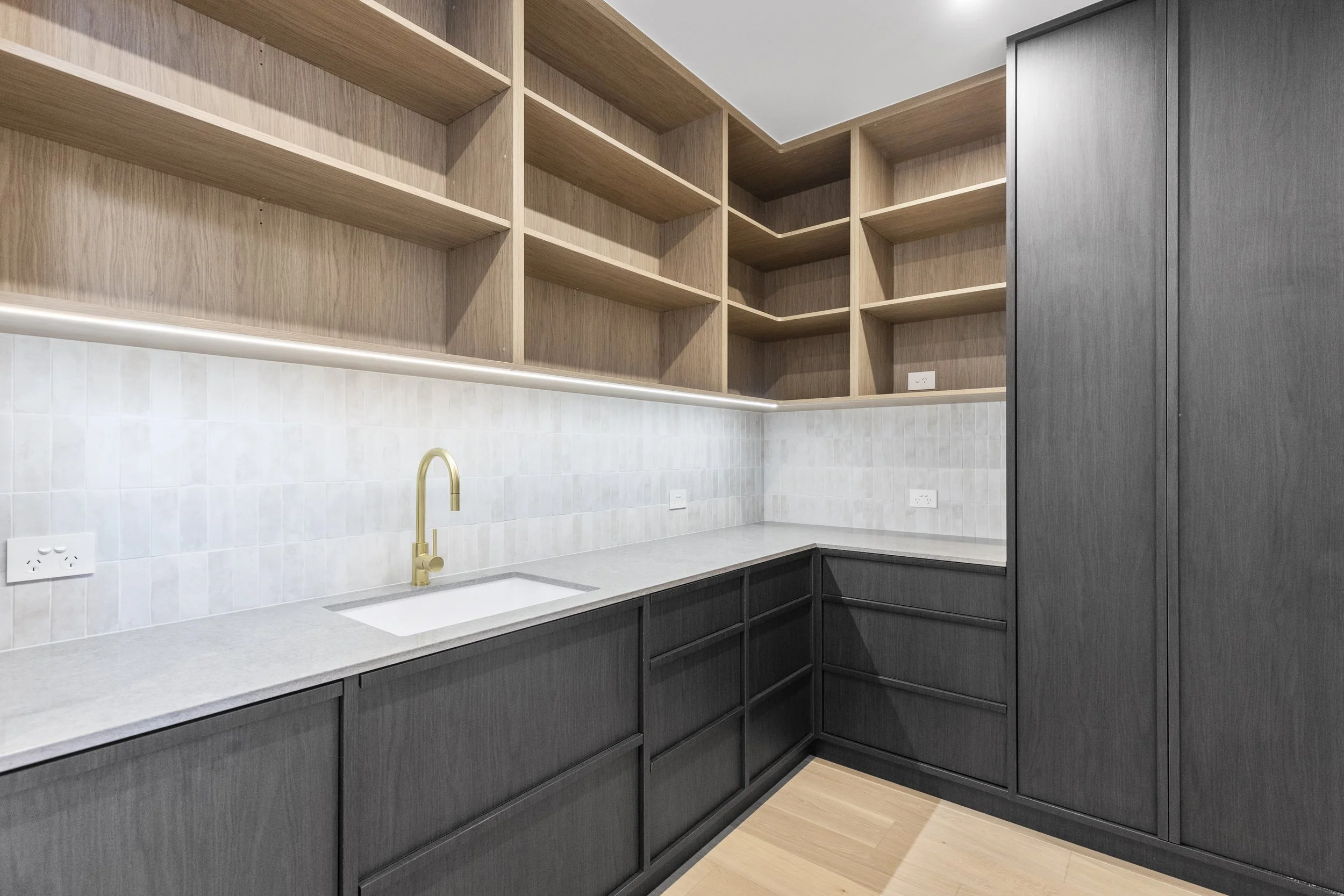 A modern kitchen with black lower cabinets, natural wood open shelves, a white backsplash, a gold faucet, and a light wood floor.