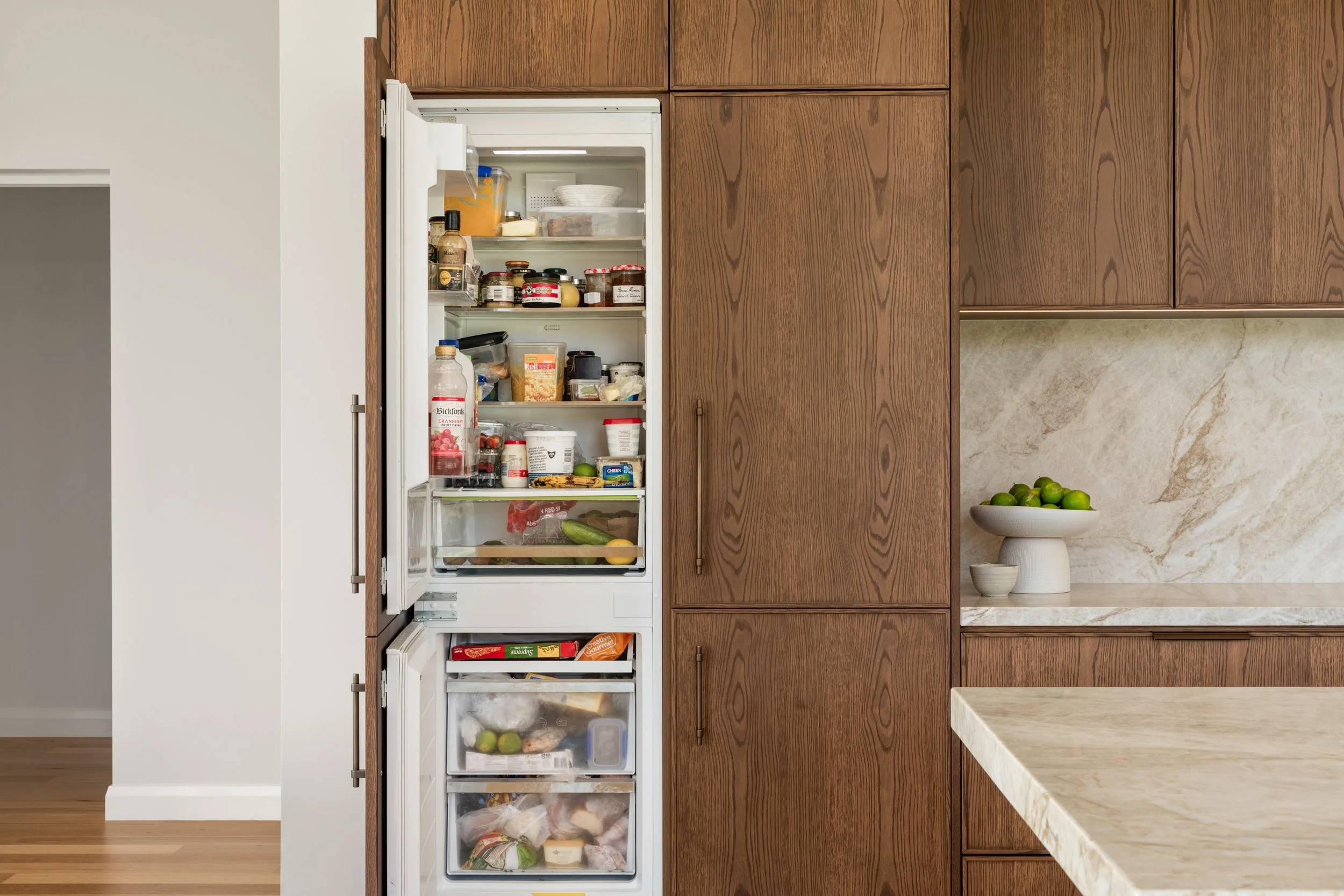 Open refrigerator revealing various food items, located next to wooden cabinets and a marble countertop with a bowl of limes.