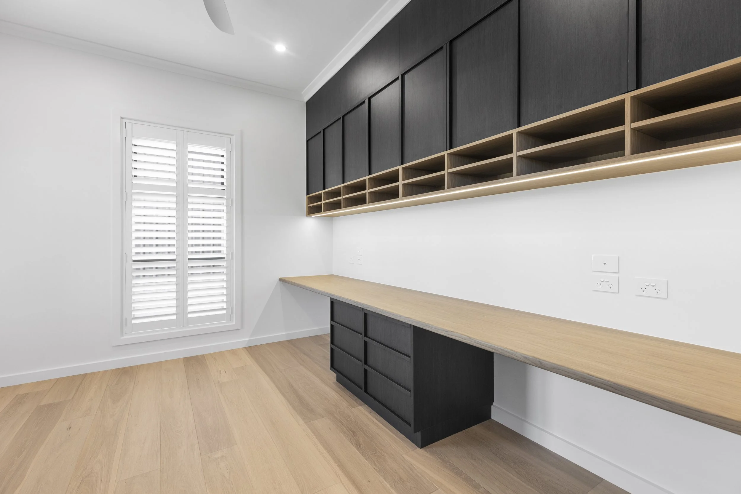 Modern minimalist kitchen with white walls, light wood flooring, black cabinets, and a wooden countertop. There is a window with white shutters and outlets on the wall.