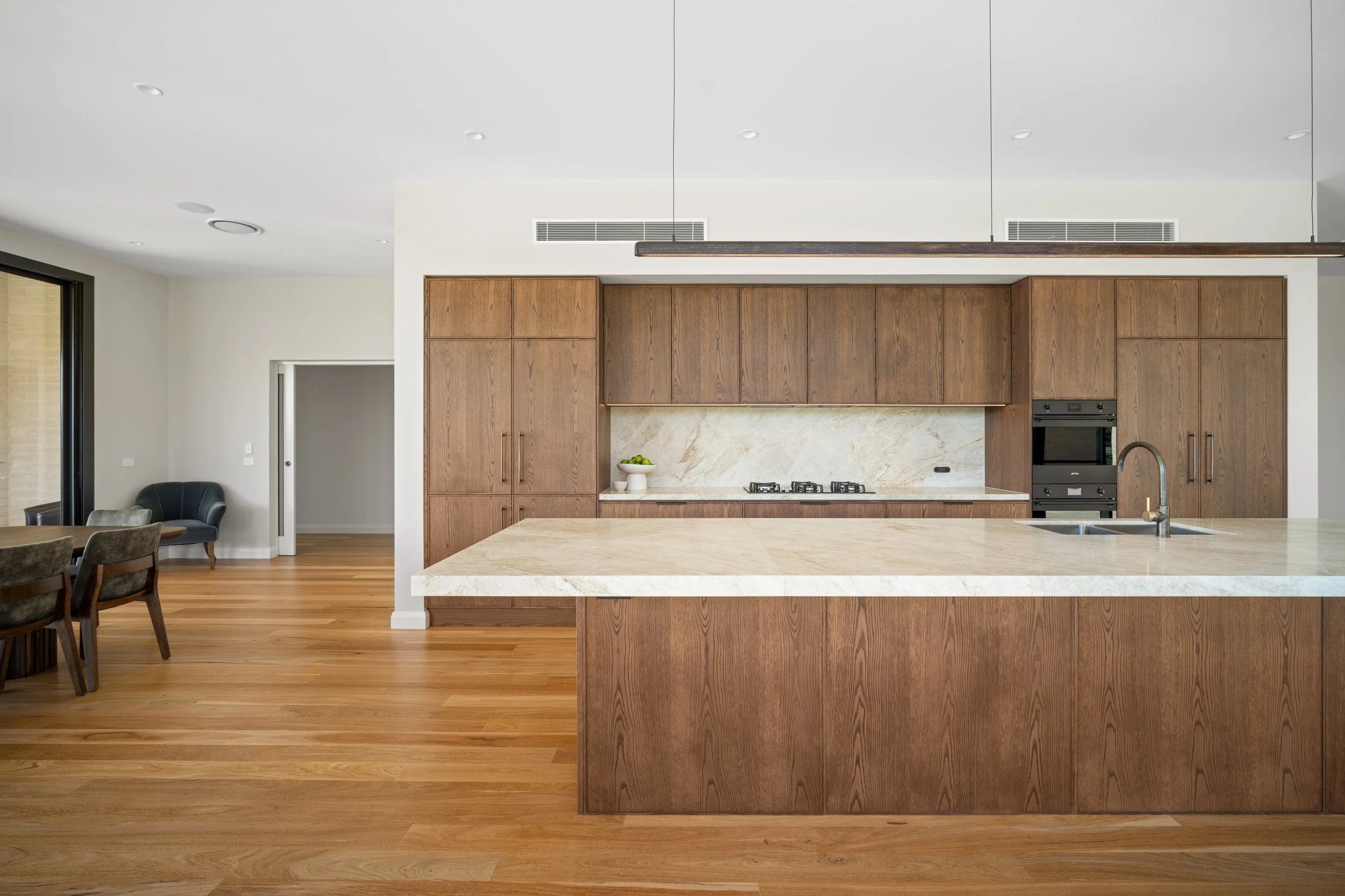 Modern kitchen with wooden cabinets, marble backsplash, granite island, and hardwood floors. A black armchair is in the adjacent living area, and sliding glass doors lead to an outdoor space.