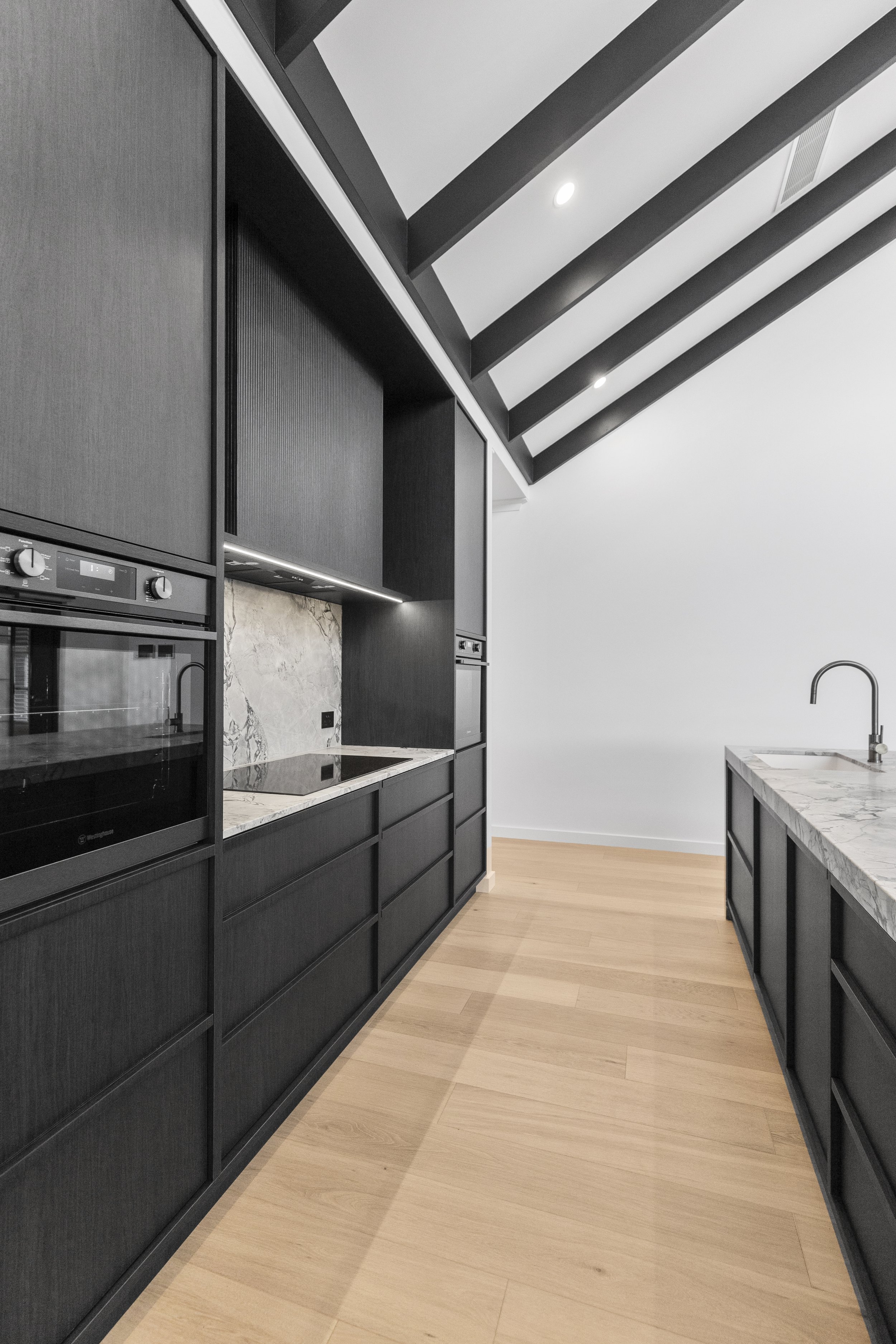 Modern kitchen with black cabinetry, marble countertops, and wooden flooring, featuring a sloped ceiling with black beams.