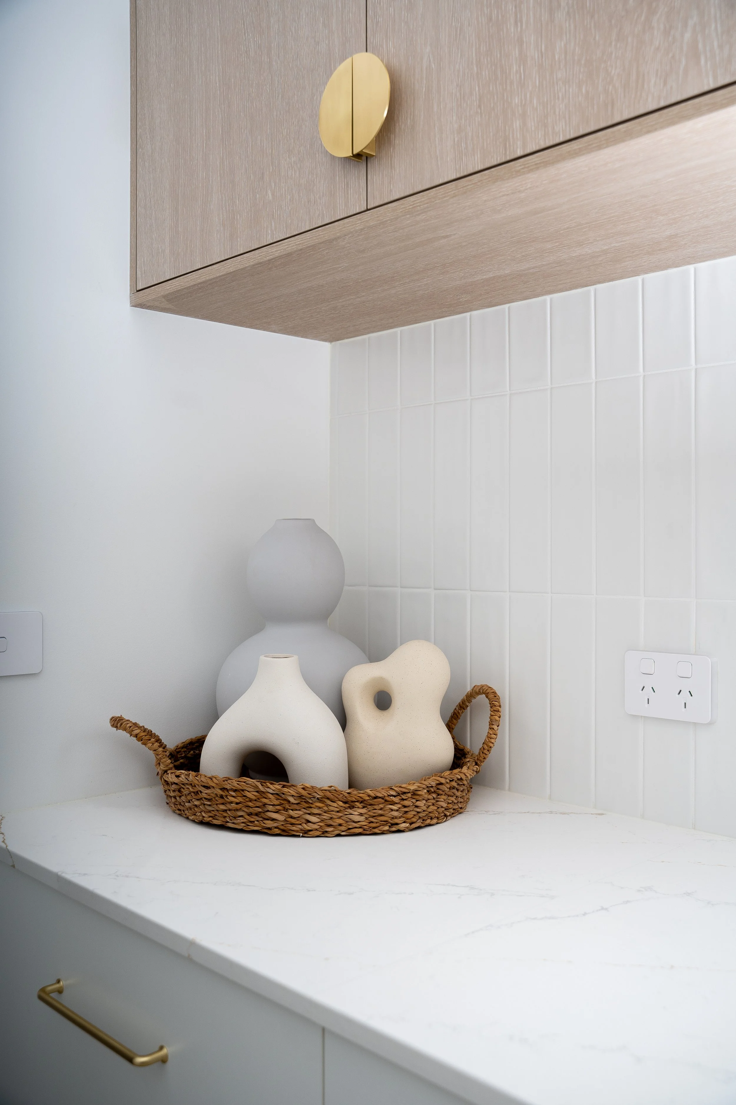 A woven basket with three beige ceramic vases of different shapes and sizes, set on a white kitchen counter beneath a wooden cabinet with a curved gold handle, and white tiled wall in the background.