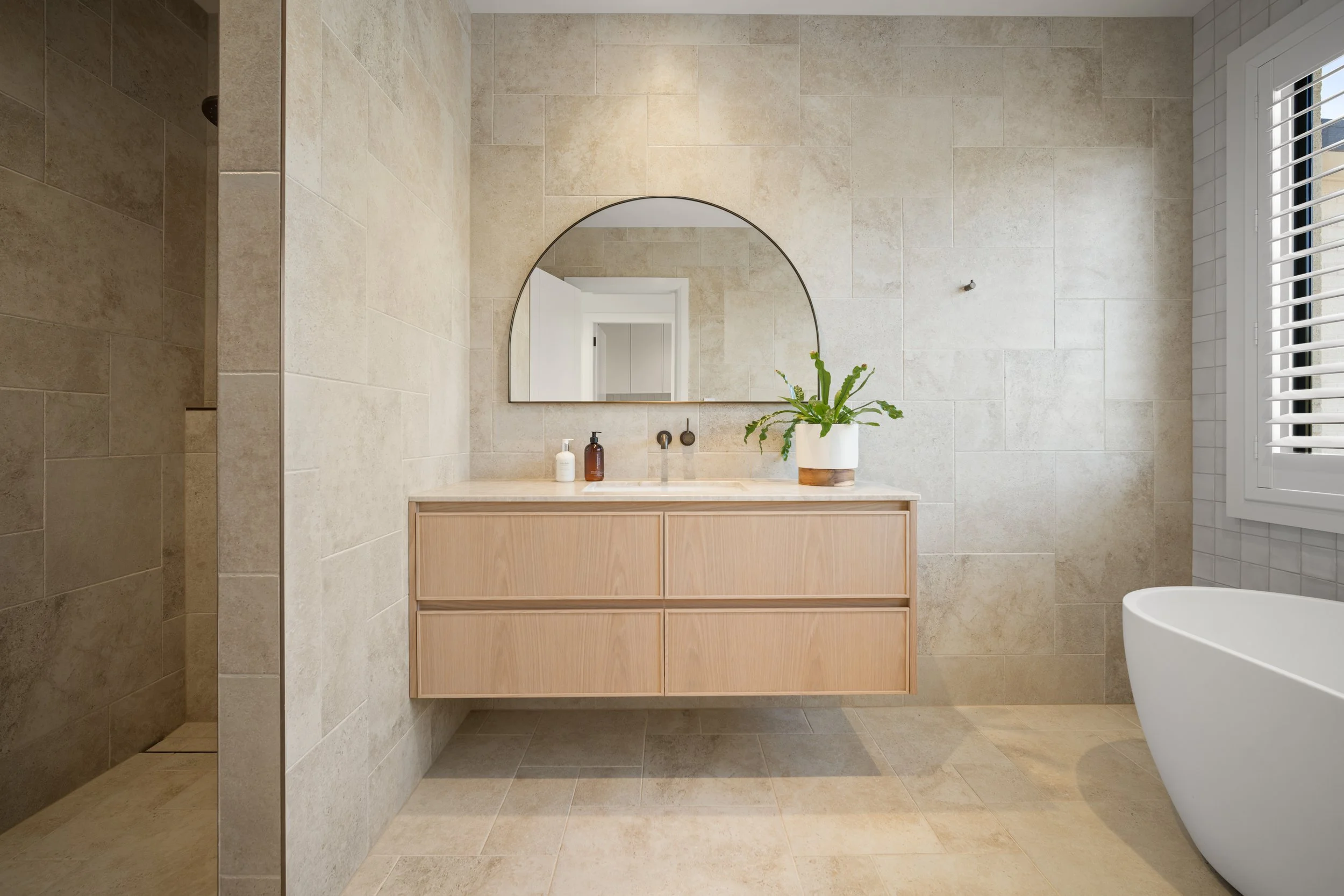 Modern bathroom with beige tiled walls and floor, a wooden vanity with a sink, a round mirror, a potted plant, and a window with white shutters.