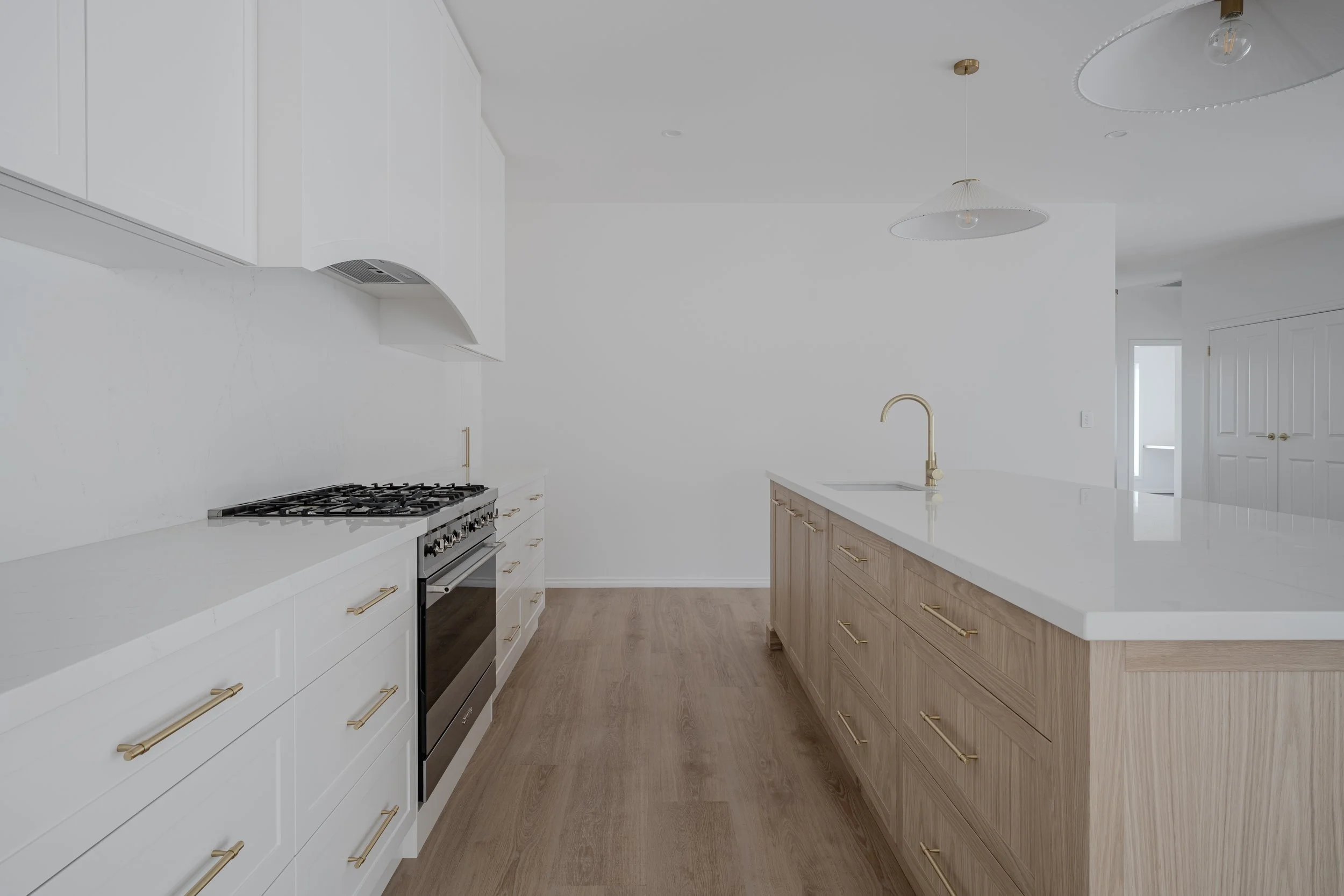 Modern kitchen with white and wooden cabinets, a white countertop, a stainless steel oven and stove, a brass faucet, and minimalist lighting fixtures.