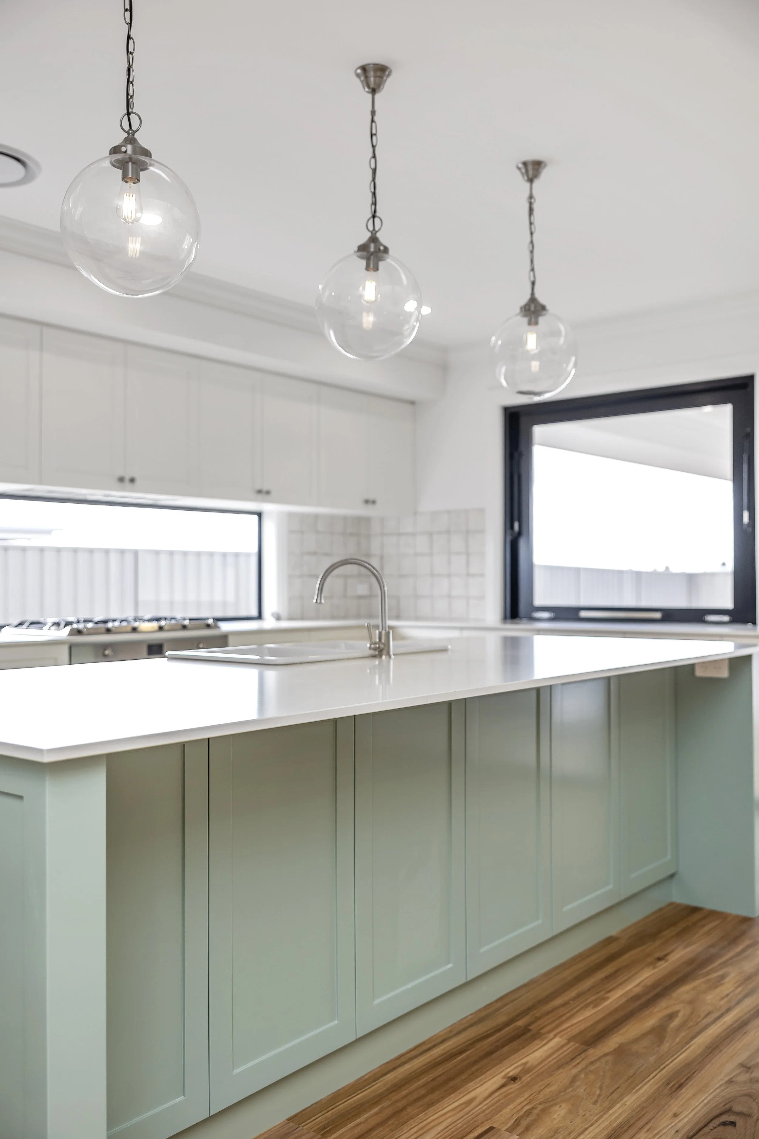 Modern kitchen with white cabinetry, a large island with light green lower cabinets, a sink, and three pendant lights hanging from the ceiling. There is a large window and hardwood flooring.