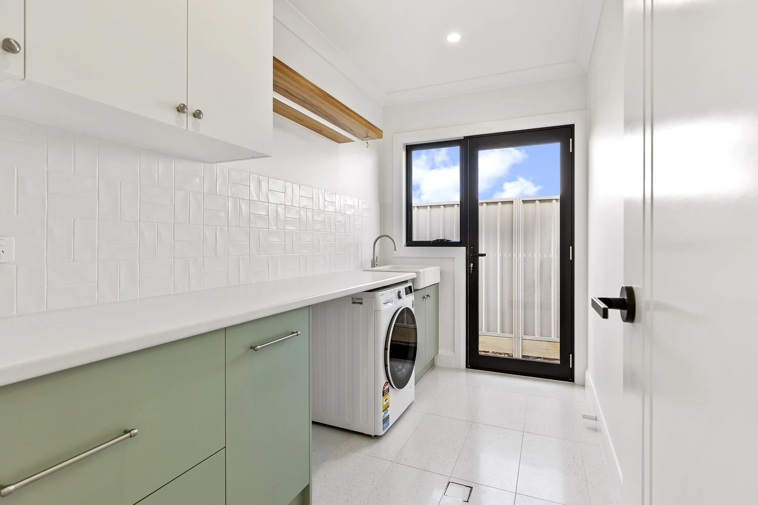 Laundry room with green cabinets, white tiled walls, a washing machine, a sink, a window showing a blue sky with clouds, and a door leading outside.