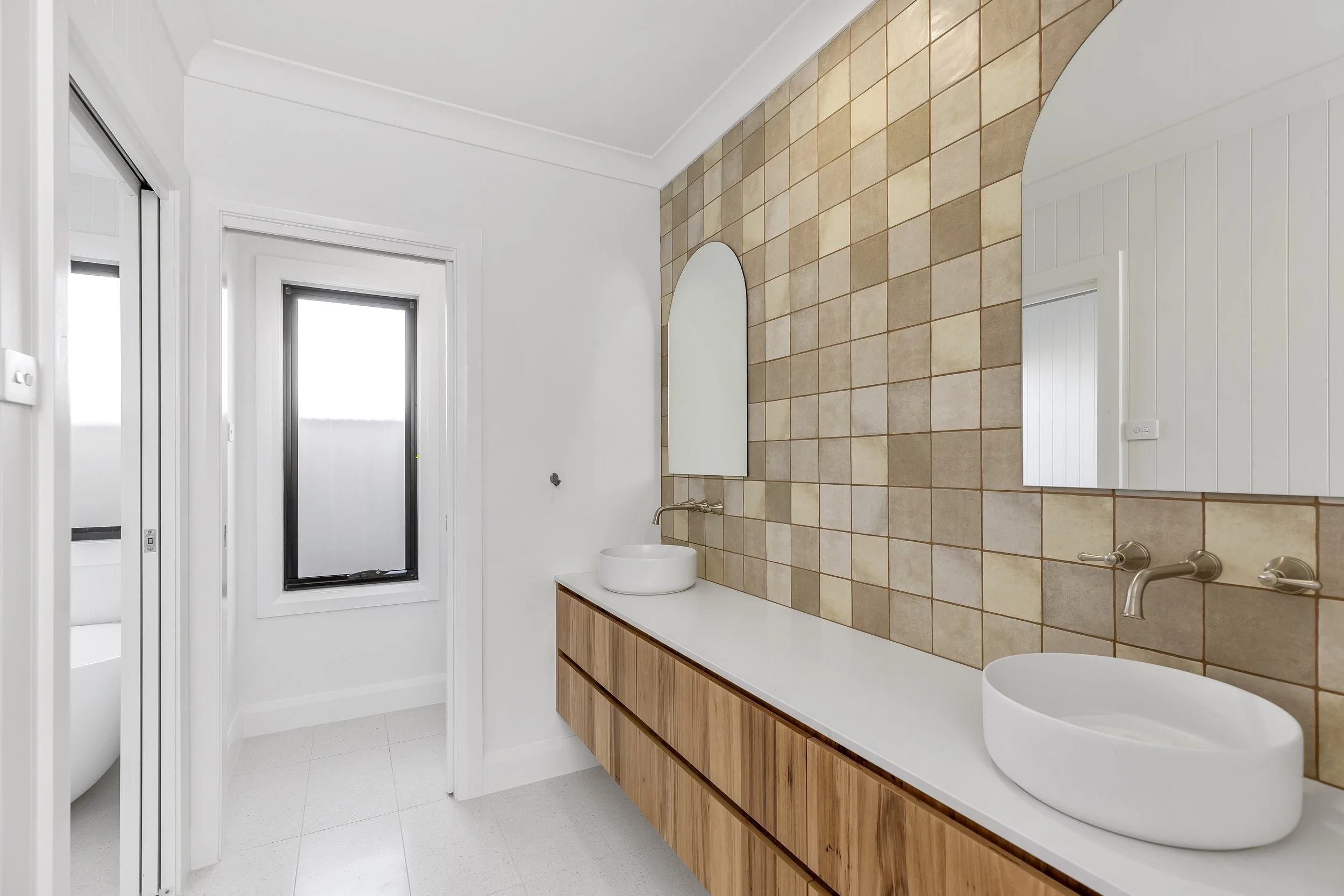 Modern bathroom with white walls, large window, beige tiled wall with mirrors, wooden vanity with vessels sinks, plus two wall-mounted faucets.
