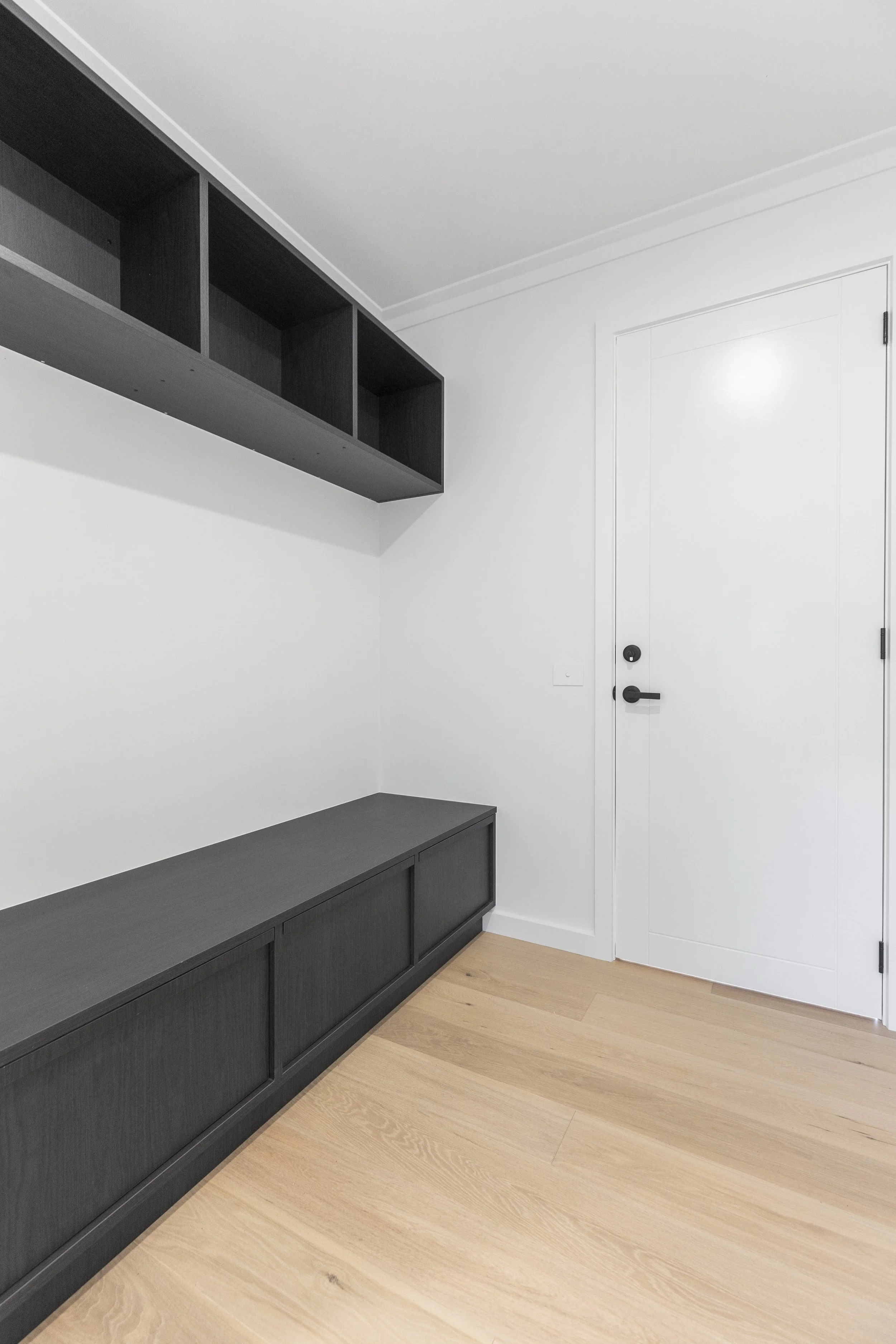 A minimalist room with white walls, light wood flooring, black storage cabinets along the wall, a black wall-mounted shelf, and a white door with black hardware.