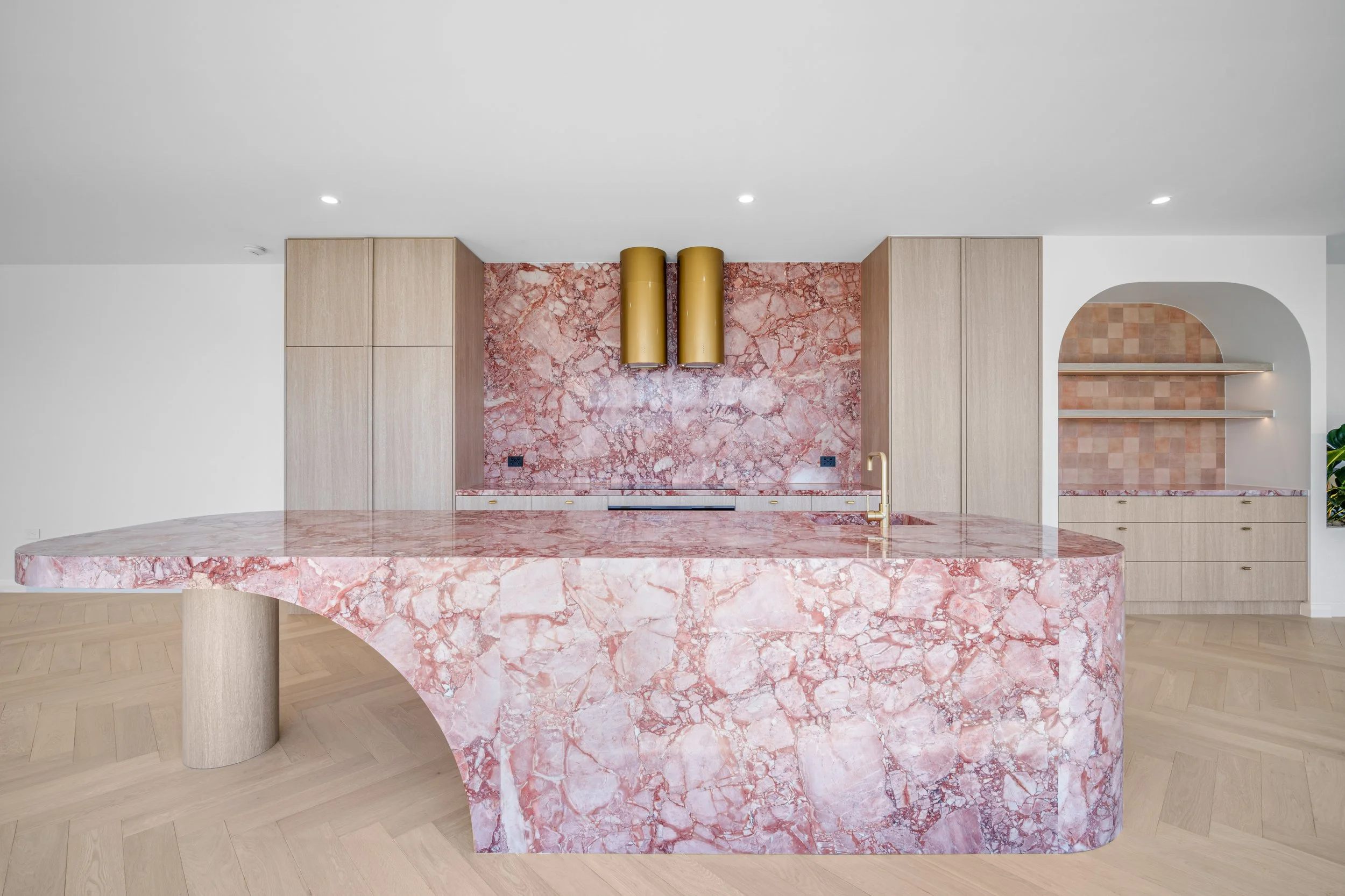 Modern kitchen with pink marble island, built-in cabinets, and a pink marble backsplash with gold cylindrical wall fixtures.