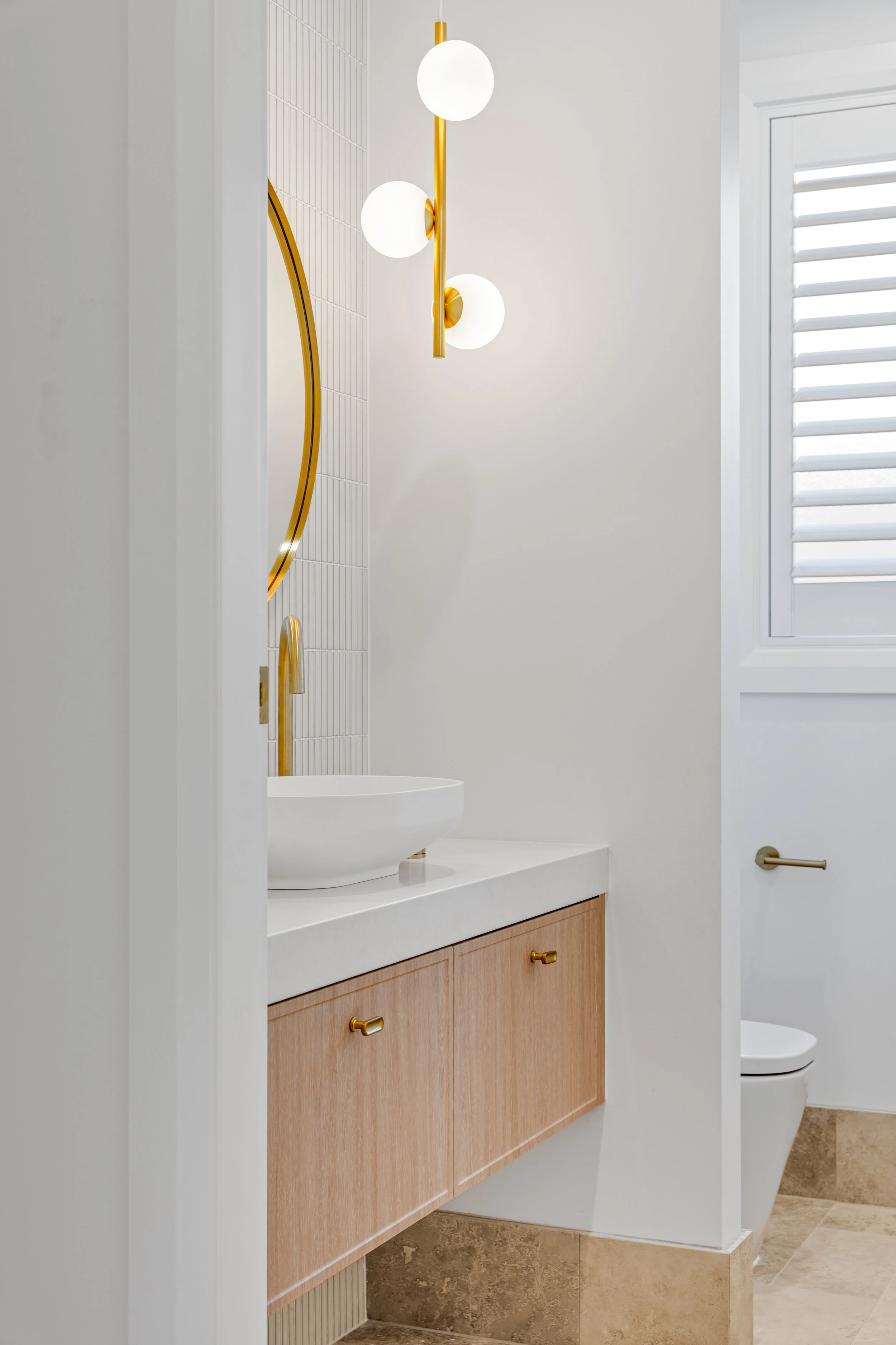 Modern bathroom with white walls, a white countertop, light wood cabinets with gold handles, a white vessel sink, a gold faucet, and a wall-mounted light fixture with three round white globes. There is a window with white blinds, and a partial view o