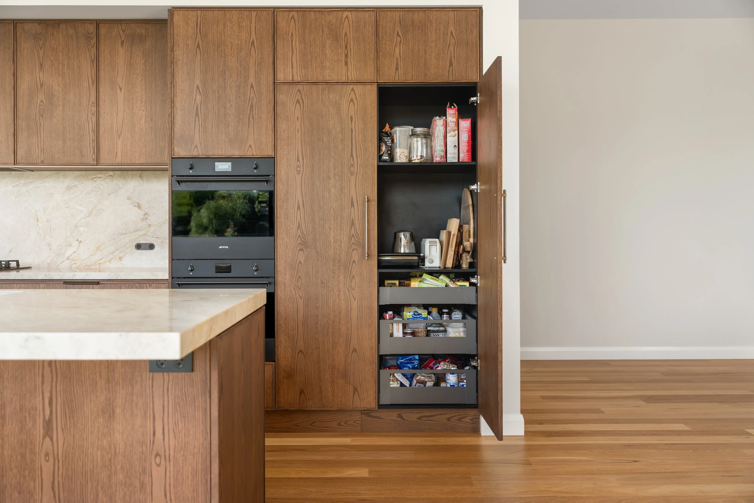 View of a kitchen with wooden cabinets, a built-in oven, and a pantry door open revealing food and kitchen supplies inside.