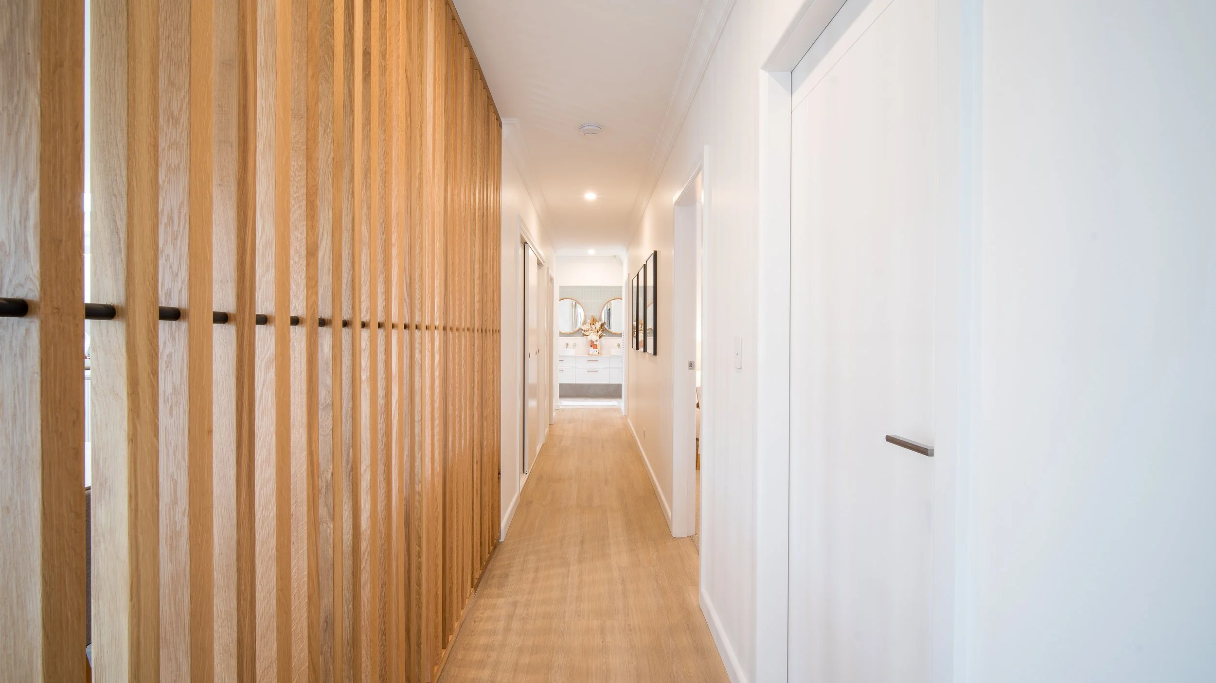 A hallway with a wooden slat divider on the left and white walls on the right, leading to a bathroom with a double vanity and mirror at the end.
