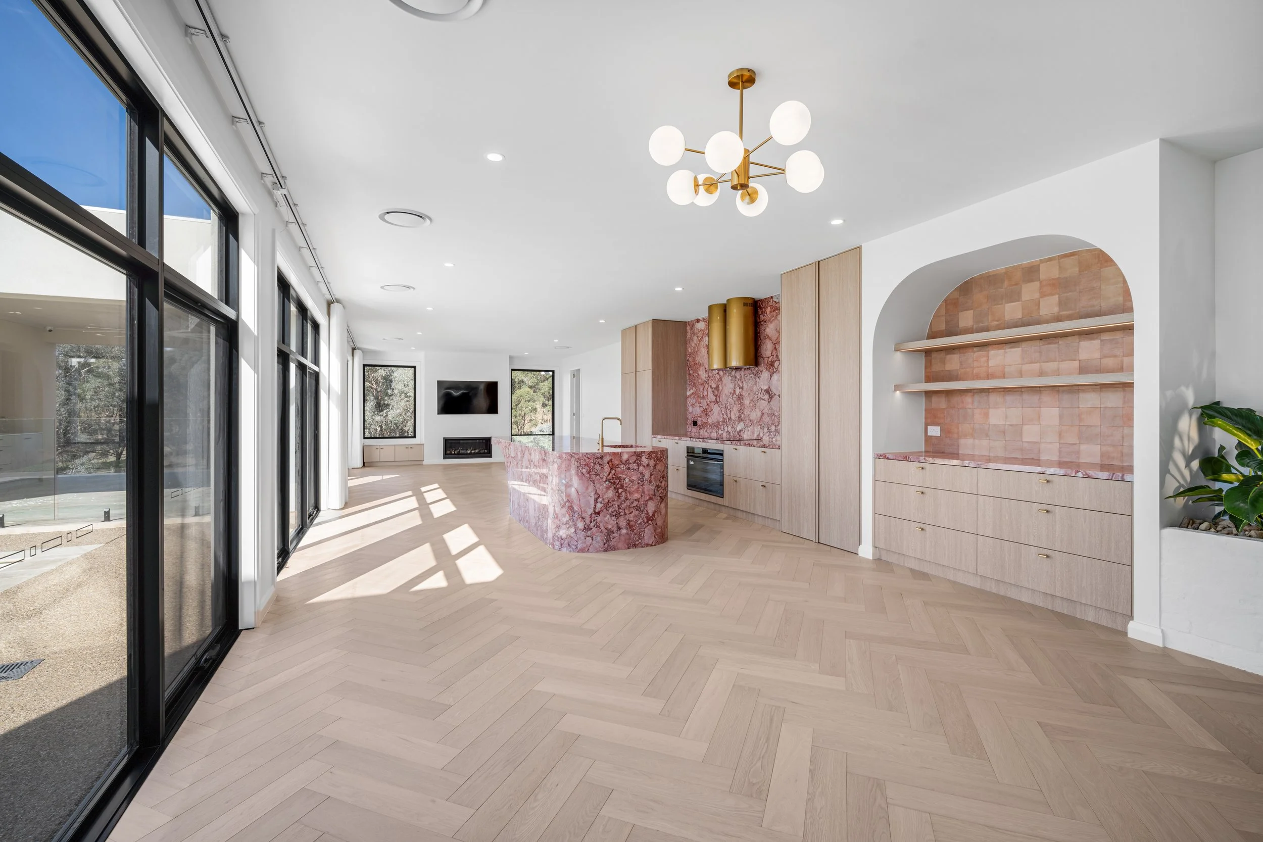 Interior of a modern house with large glass sliding doors, light wood flooring, a kitchen with pink marble island, and minimal decor.