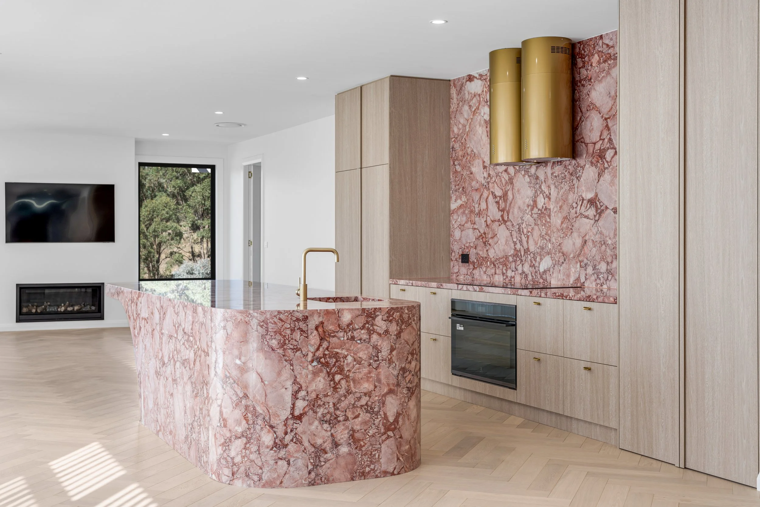 Modern kitchen with pink marble island and matching backsplash, light wood cabinetry, gold range hood, and a window showing trees outside.