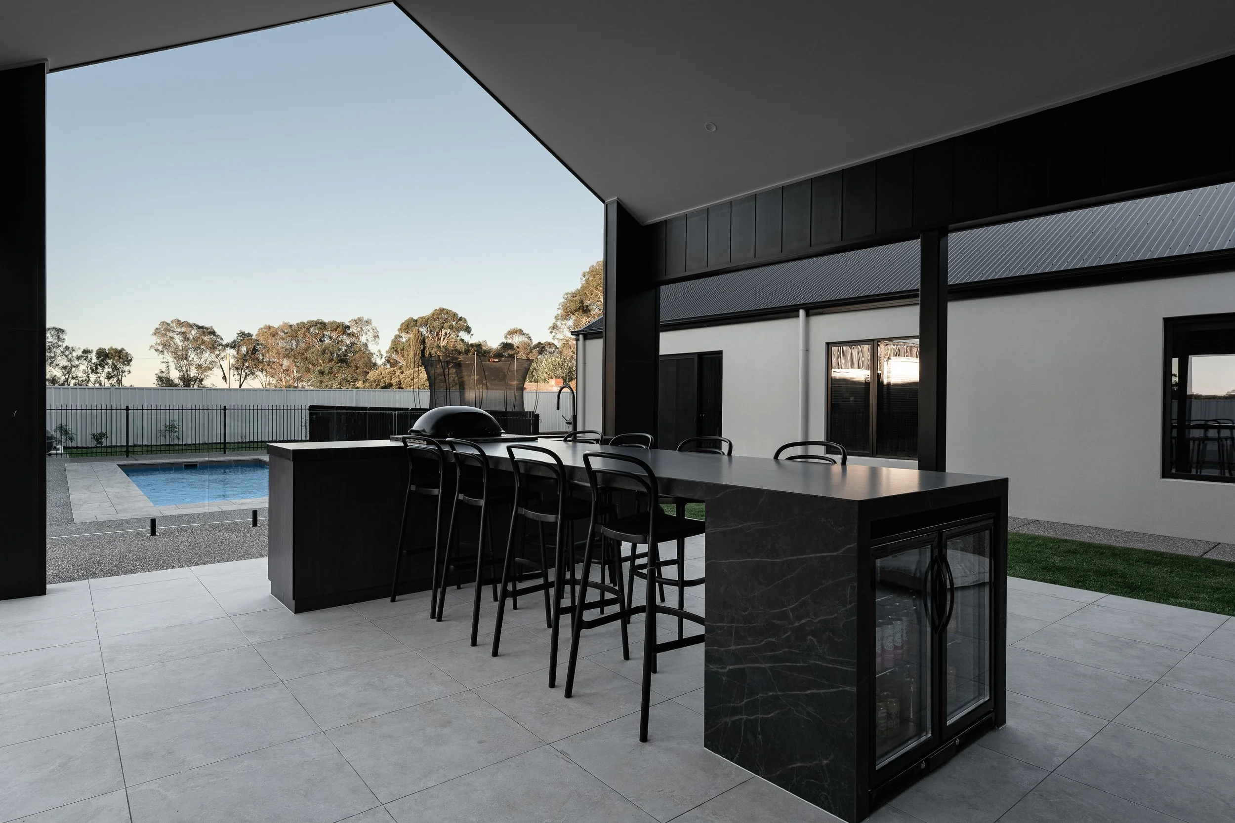 Modern outdoor patio area with a long black dining table, black chairs, a pool, trampoline, and trees in the background under a clear sky.