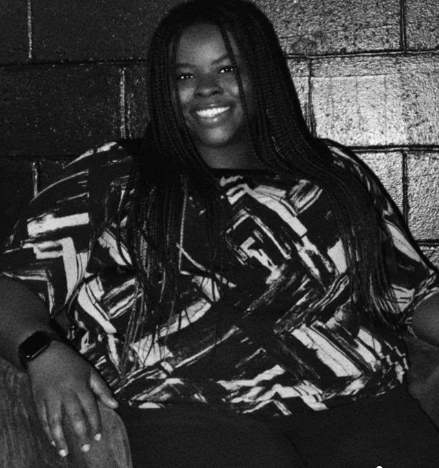 A woman with braided hair smiling, sitting against a brick wall, wearing a patterned top and a watch.