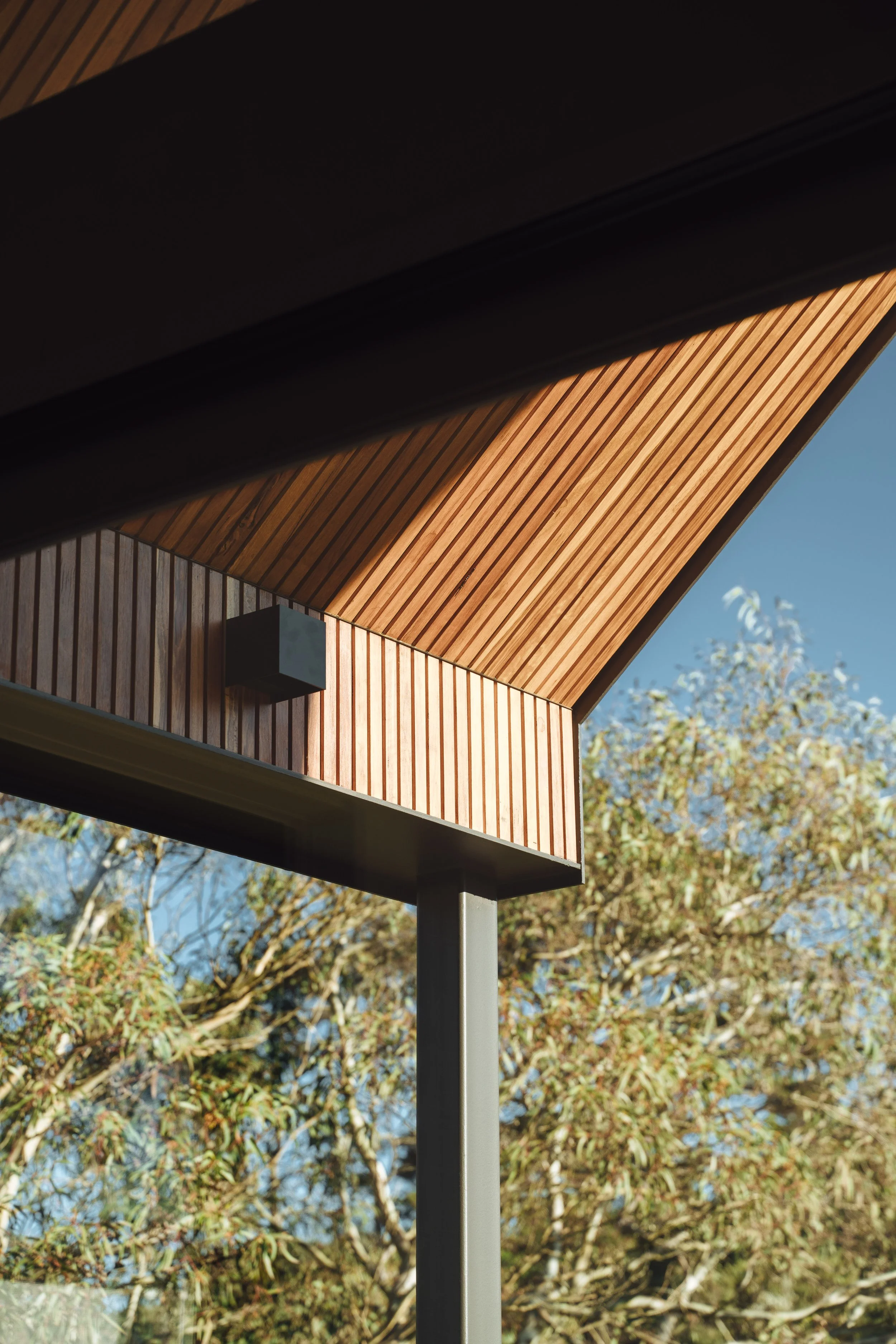 A modern birdhouse made of wood with a sloped roof, mounted on a metal pole against a background of trees and a clear blue sky.