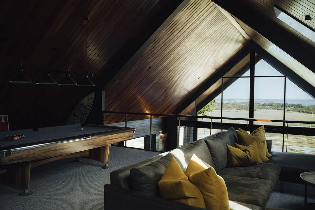 Interior of a loft with a sloped wooden ceiling, a pool table, and a large window overlooking a landscape.