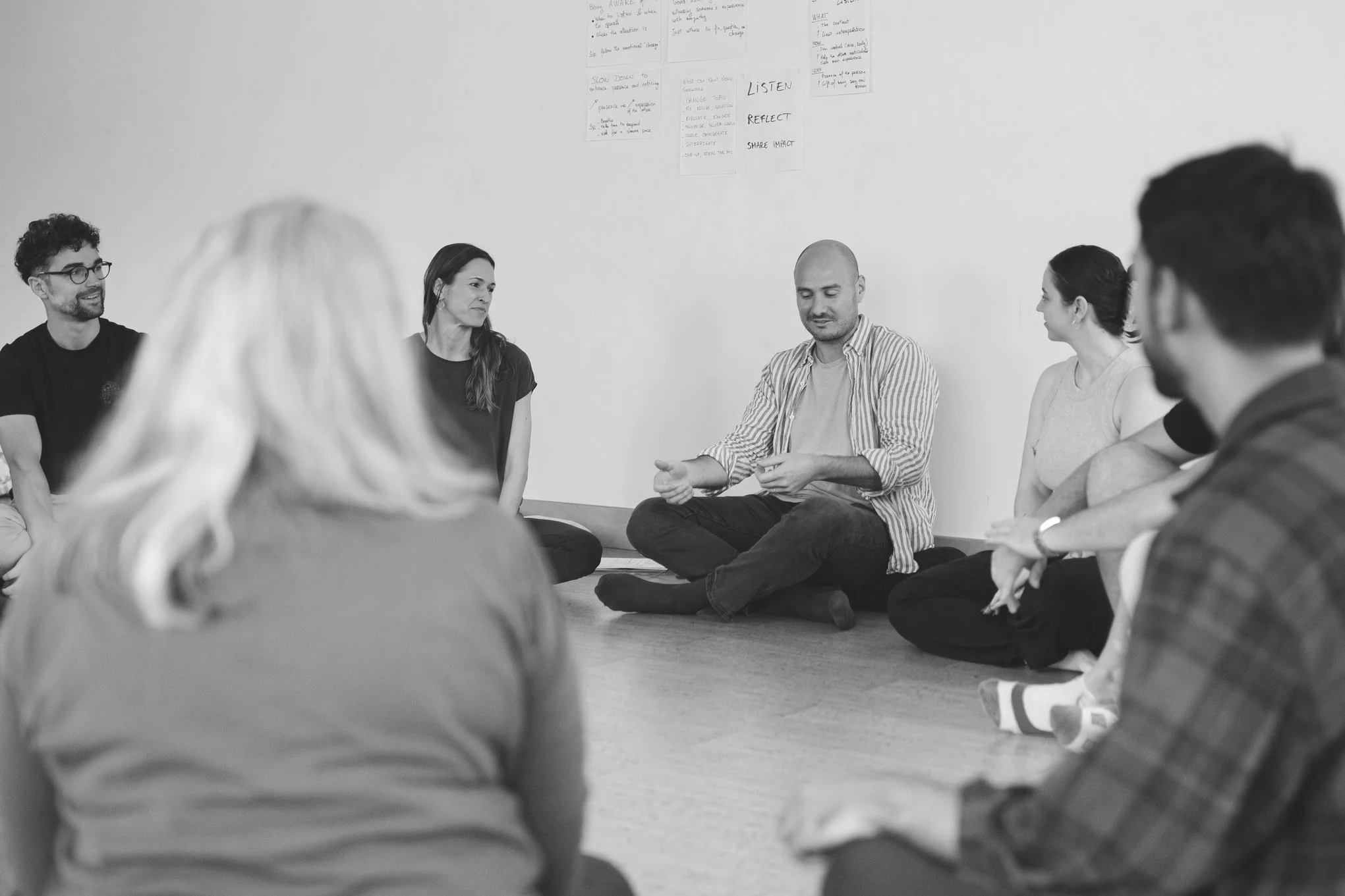 People sitting cross-legged on the floor in a circle during a group discussion in a room with notes on the wall.