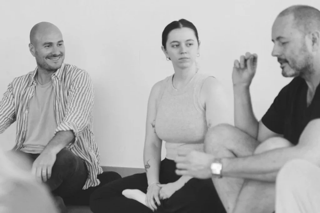 Three people sitting on the floor in a circle, engaging in conversation. The man on the left wears a striped shirt, the woman in the middle wears a sleeveless top, and the man on the right wears a dark T-shirt and is gesturing with his hand.