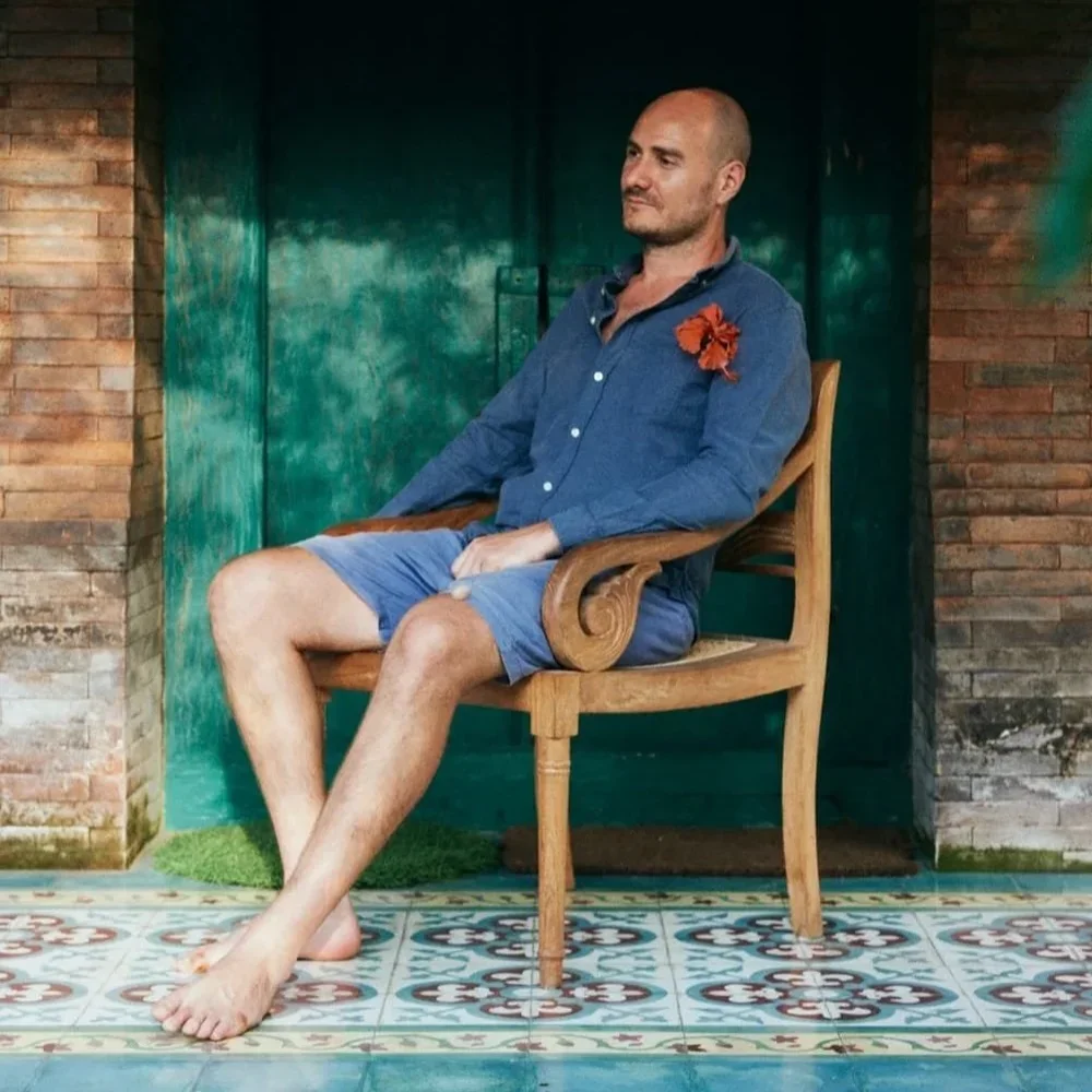 A man with a shaved head sitting barefoot on a wooden chair in a room with brick walls and colorful tiled floor, wearing a blue shirt and shorts, with an orange flower pinned to his shirt.