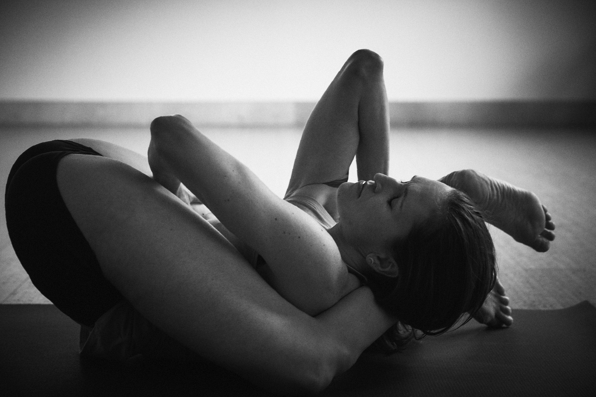 Amanda Ferris lying on the floor in a yoga pose with their arms extended and legs bent, in a black-and-white photograph.