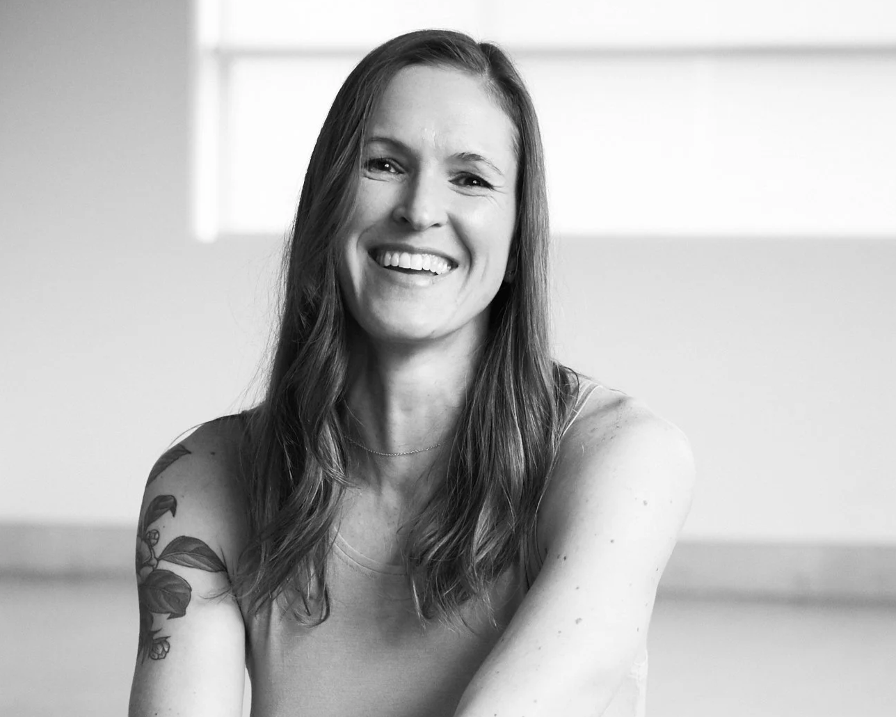 Black and white photo of Amanda Ferris with long wavy hair, tattoo on her left arm, wearing a sleeveless top, sitting with arms crossed in a bright room.