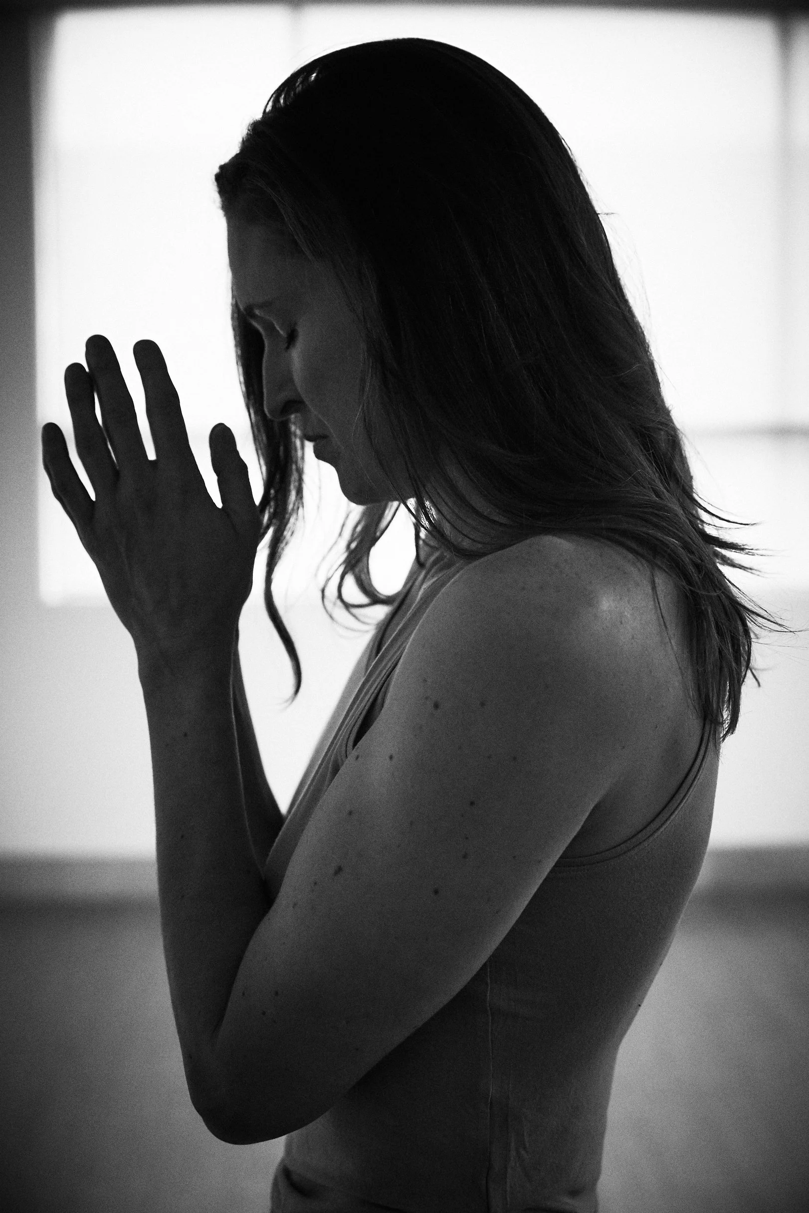 A black and white photo of a woman with long hair and freckles, her head bowed and her hands together in a praying position.