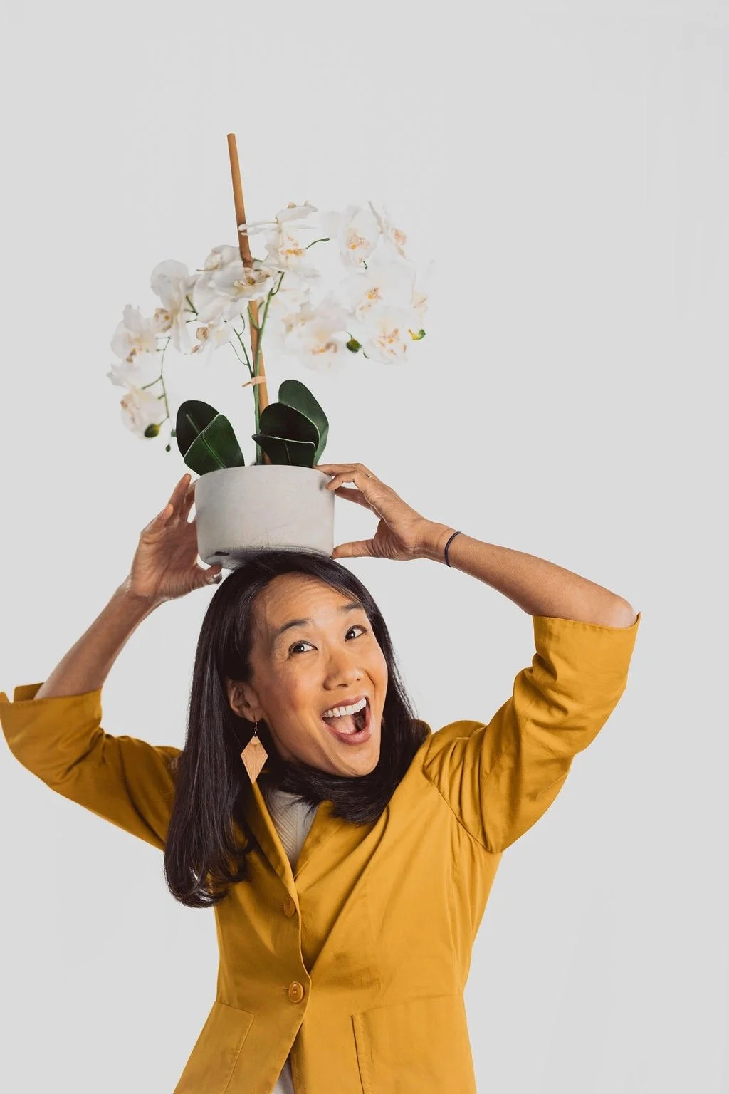 A woman in a mustard-colored jacket holding a potted white orchid plant above her head and smiling.