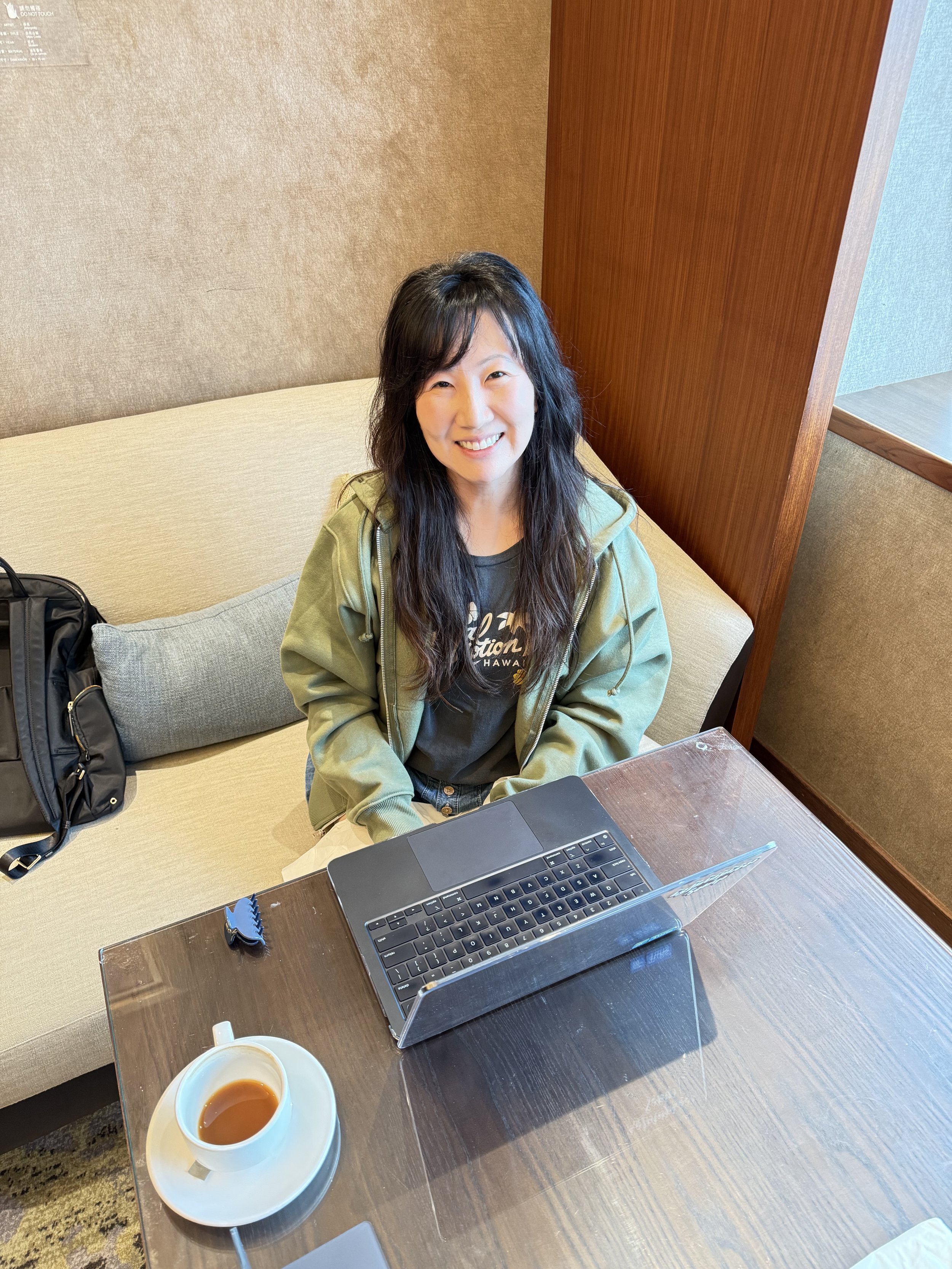 A woman with long dark hair sitting at a table with a MacBook laptop, a cup of tea, a black backpack, and a small blue whistle, in a room with brown and beige walls and wooden accents.