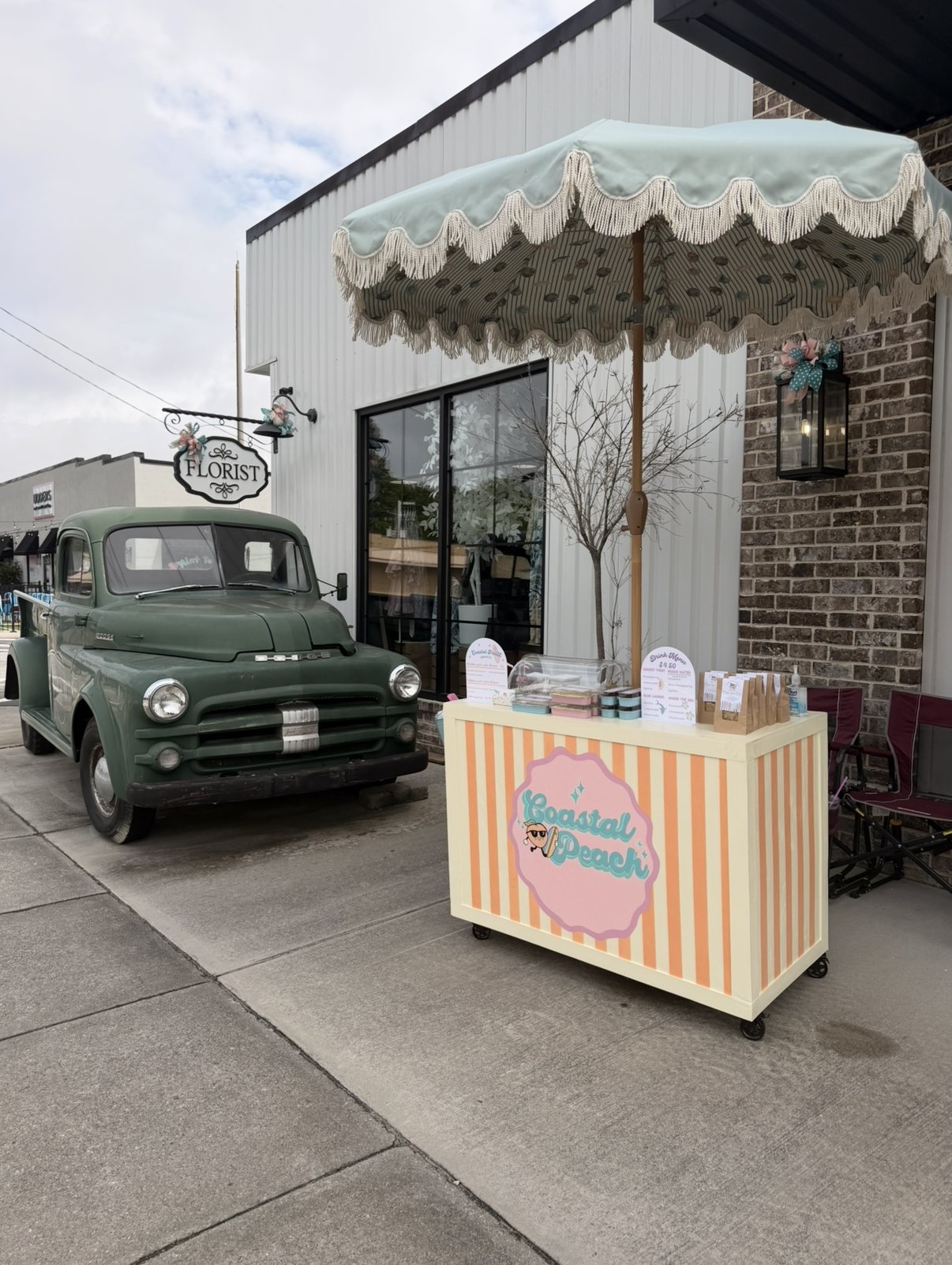 A vintage green pickup truck parked outside a storefront with a sign that reads 'Florist'. In front of the store, there is a beach-themed ice cream or dessert stand labeled 'Coastal Peach' with striped pink, yellow, and white patterns, and items like jars of toppings on top. A striped patio umbrella shades part of the stand. The store has large glass windows, a leafless tree beside it, and decorative lanterns on the brick wall.