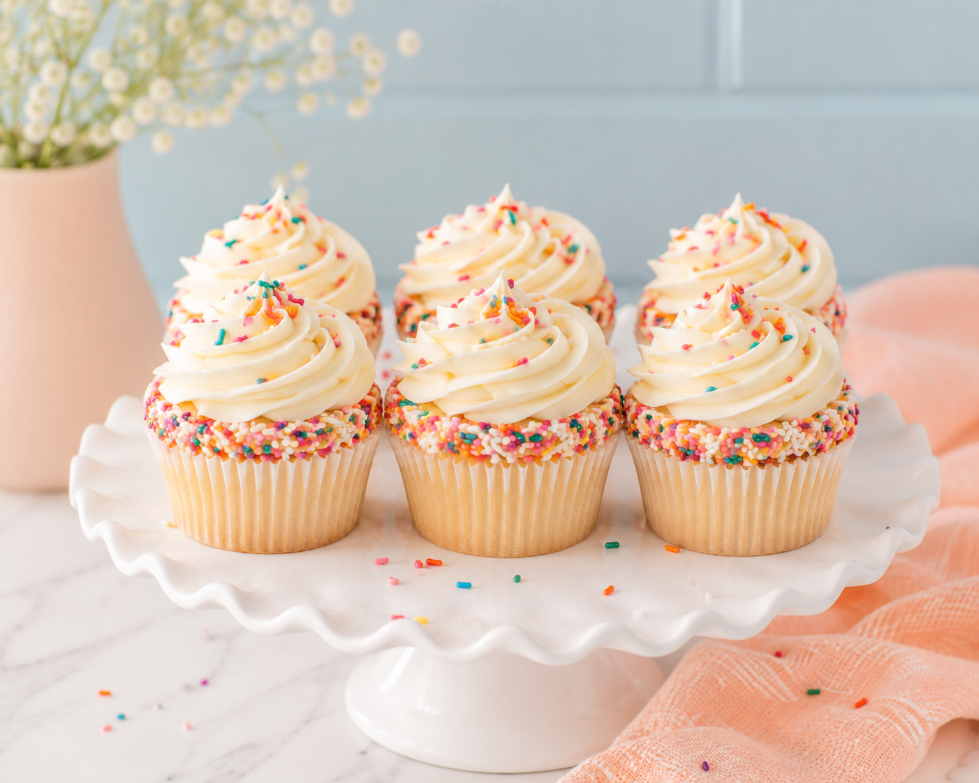 Six cupcakes with white frosting and colorful sprinkles arranged on a white scalloped cake stand, with a pink vase with white flowers in the background on the left and a pink cloth on the right.