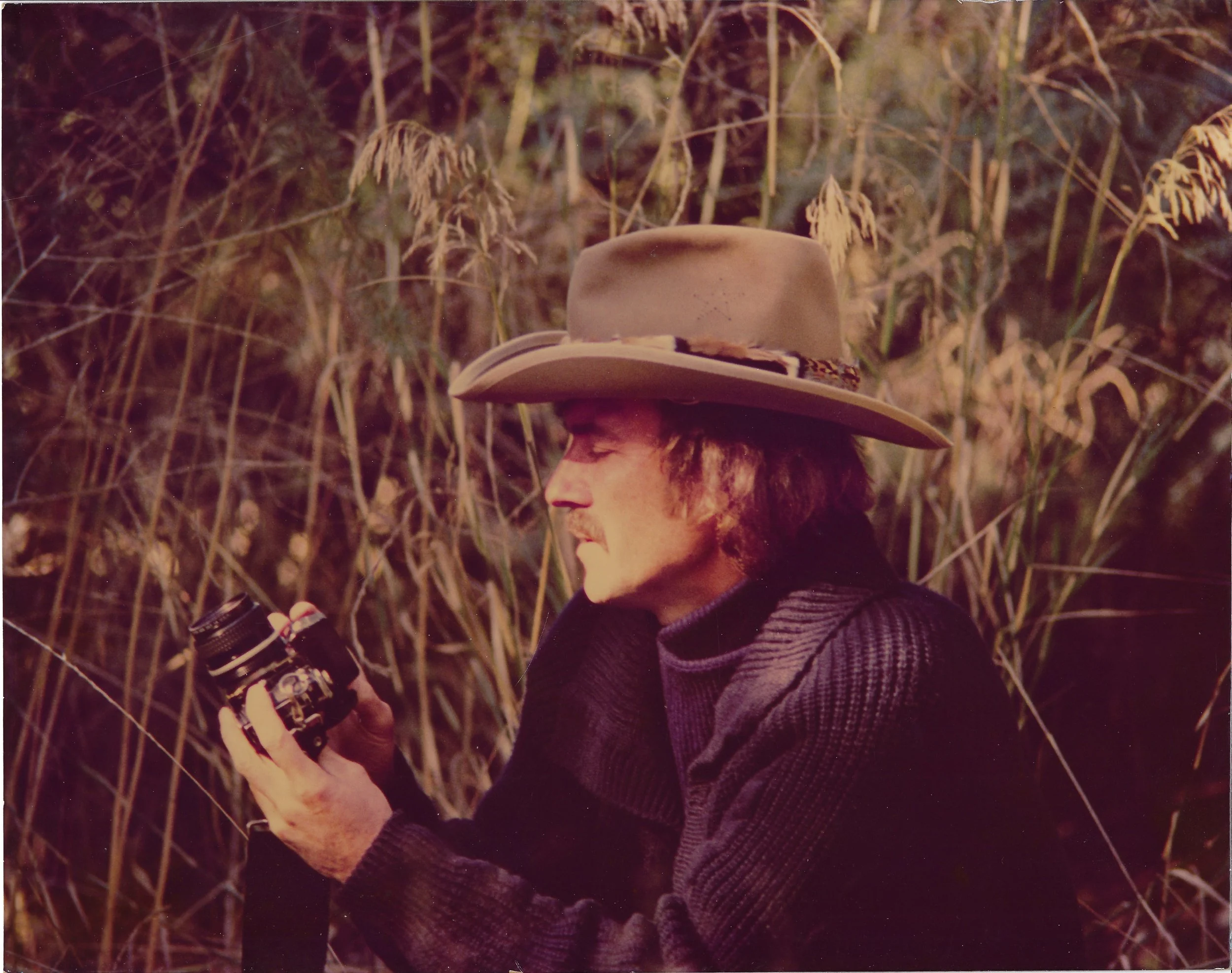 A person wearing a wide-brimmed hat and a dark jacket, standing outdoors among tall, dry grass or crops, holding a small bottle or container.
